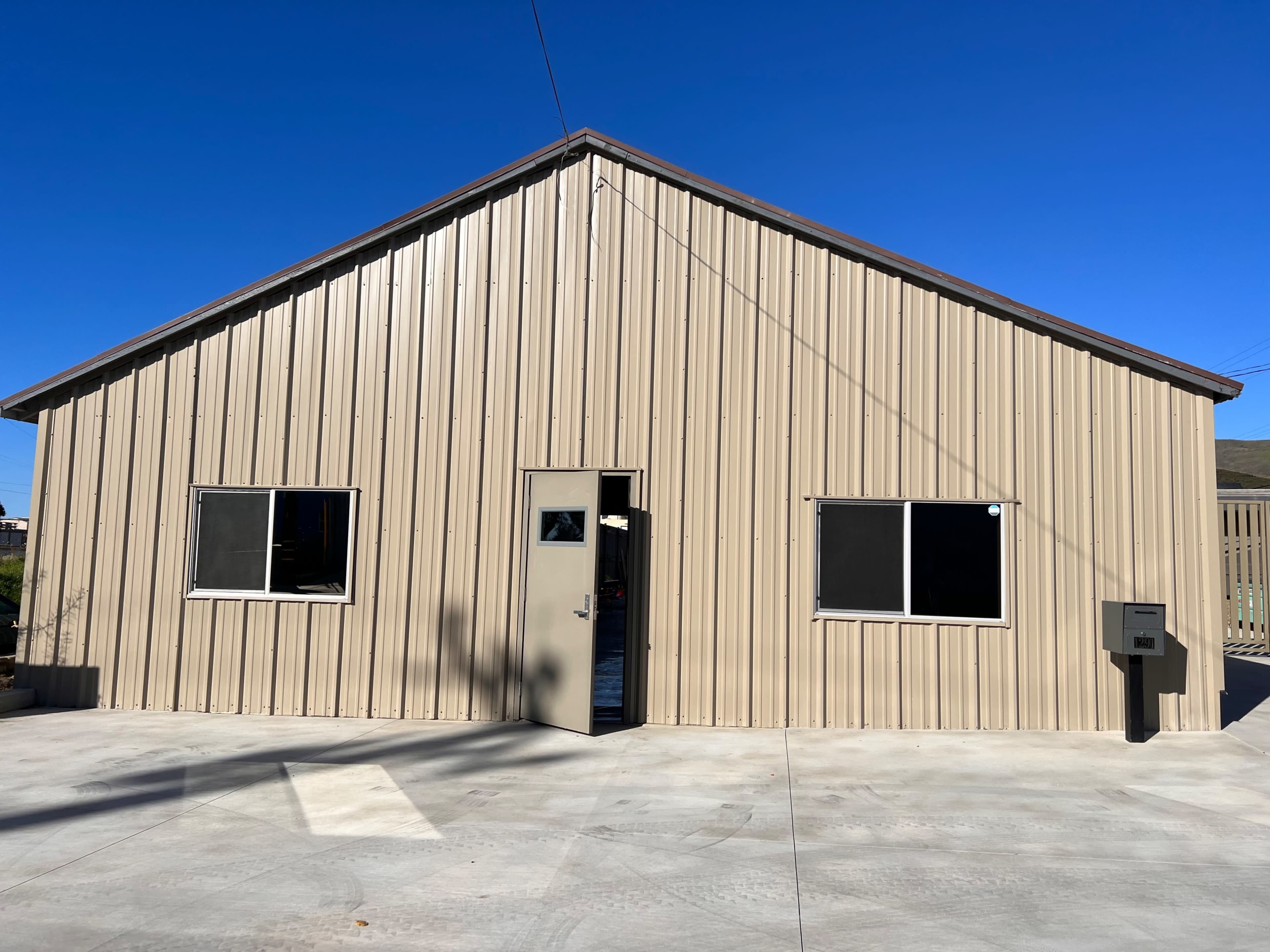 A metal-clad building with a sloped roof, two windows, and a single door is set against a clear blue sky.