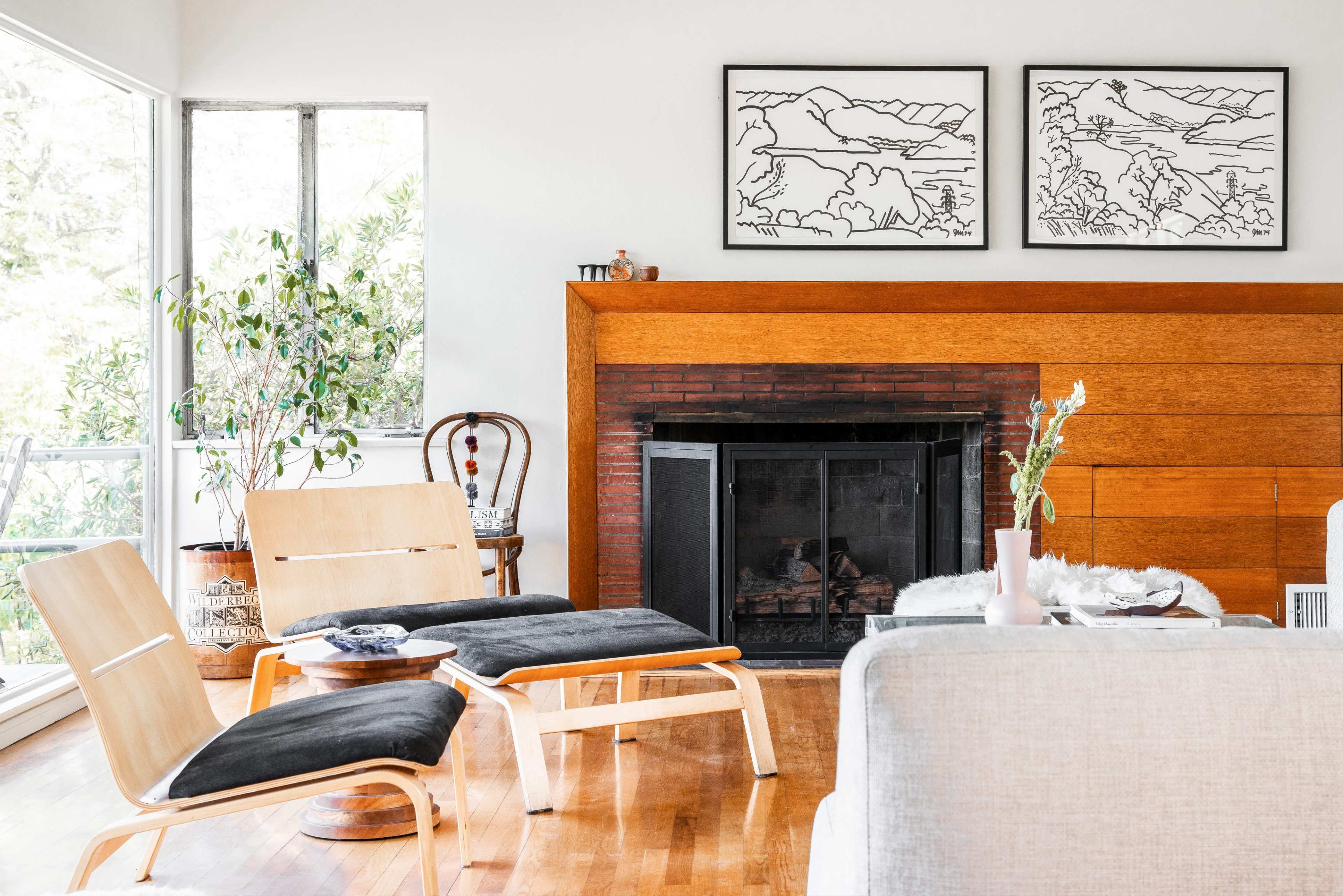 The scene features a modern living room with large windows, a wooden fireplace, framed line drawings on the wall, and minimalist furniture, including two lounge chairs and a sofa.