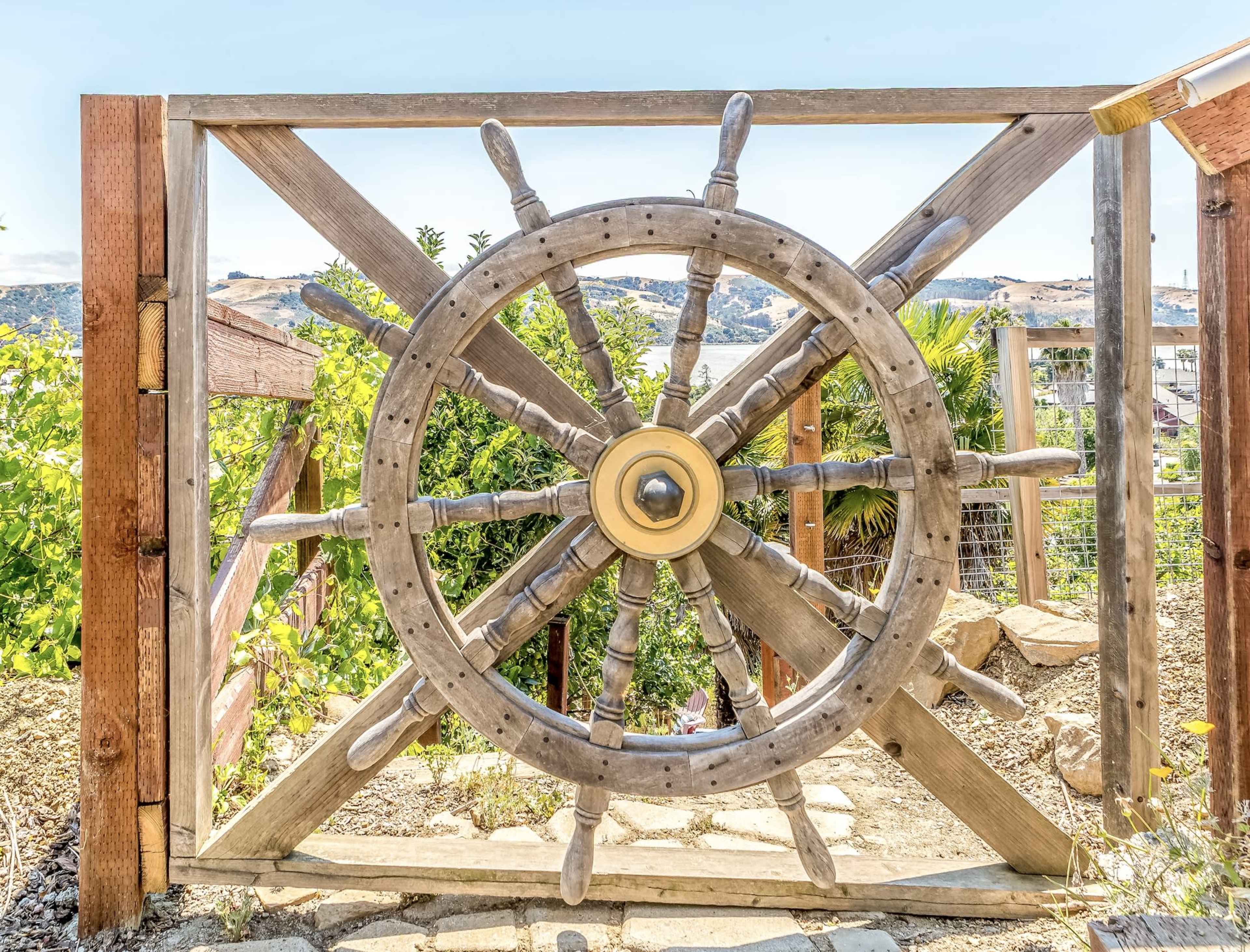 A large wooden ship's wheel serves as a gate, framed by a wooden structure and surrounded by greenery.