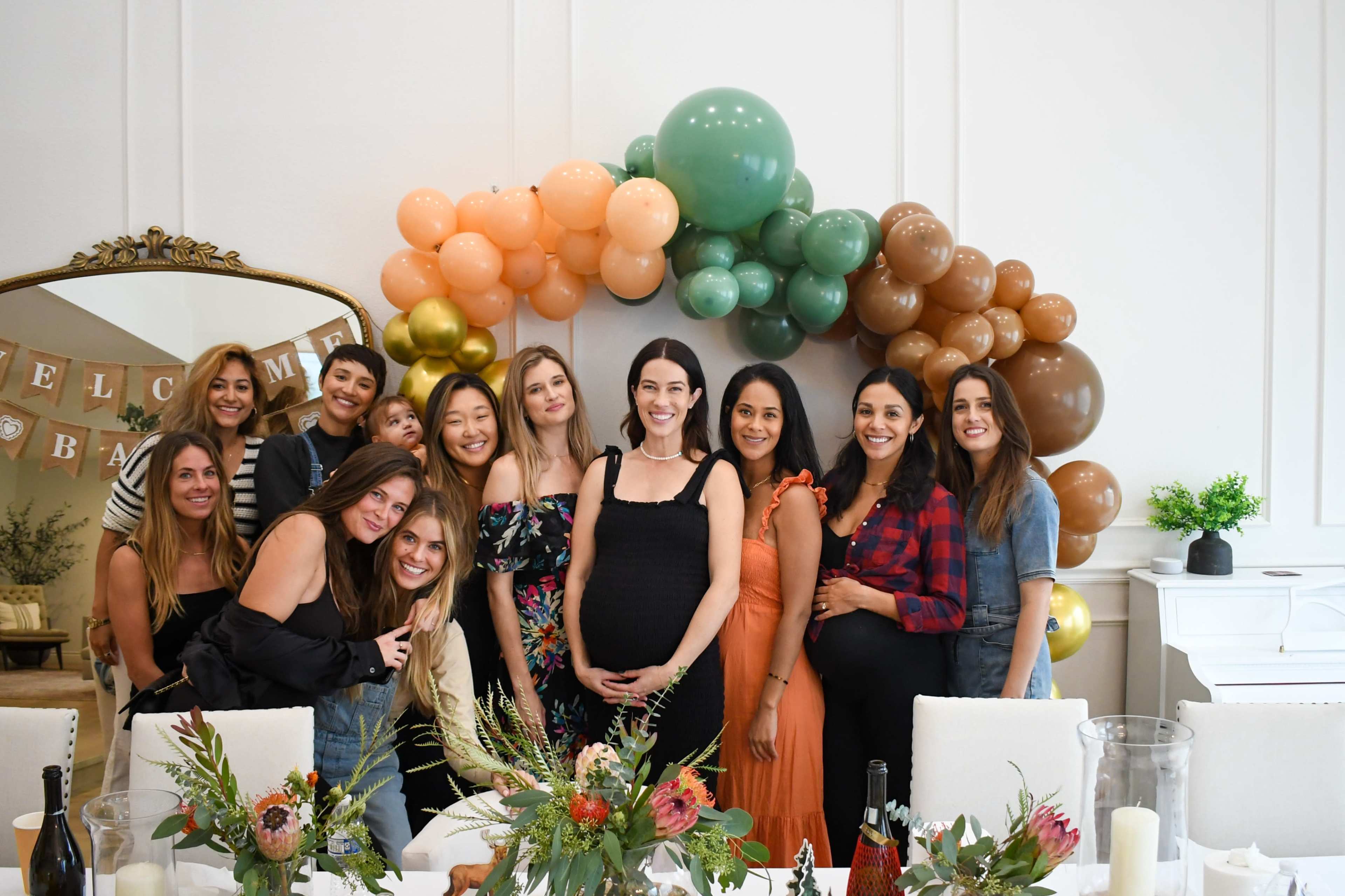 A group of twelve women stands together in a decorated room featuring a balloon arch, celebrating an event.