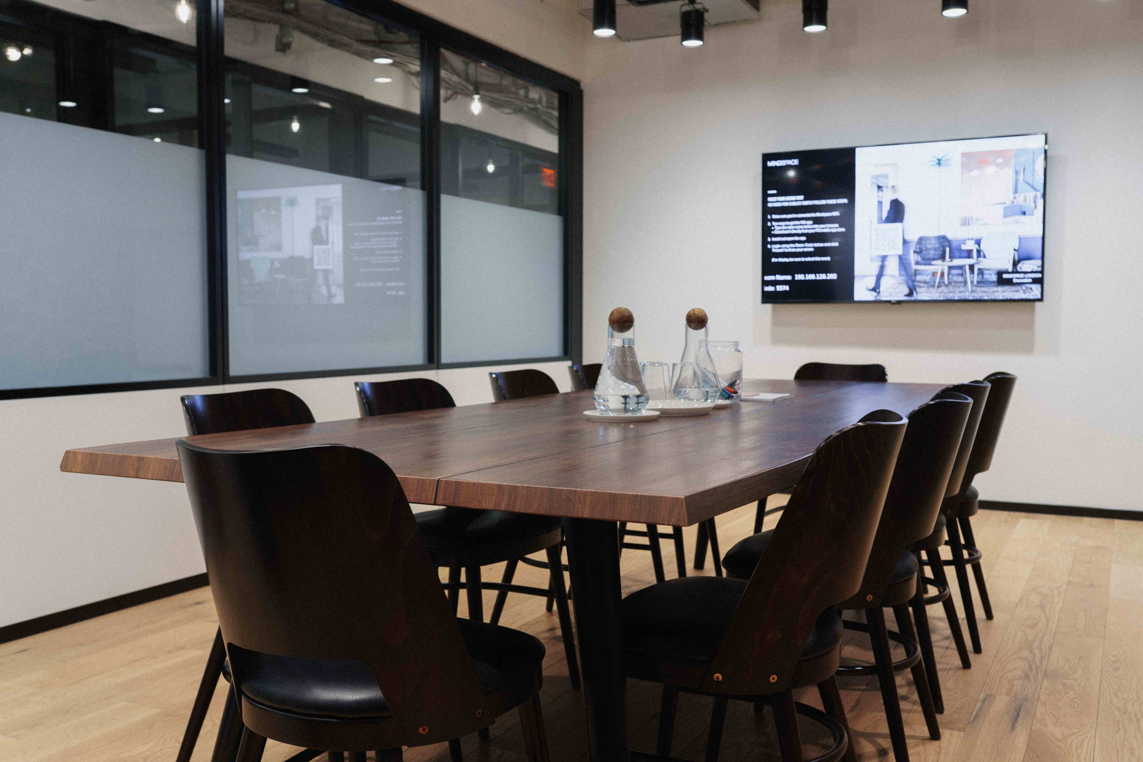 A long wooden conference table surrounded by black chairs is set in a modern meeting room with a wall-mounted screen displaying information.