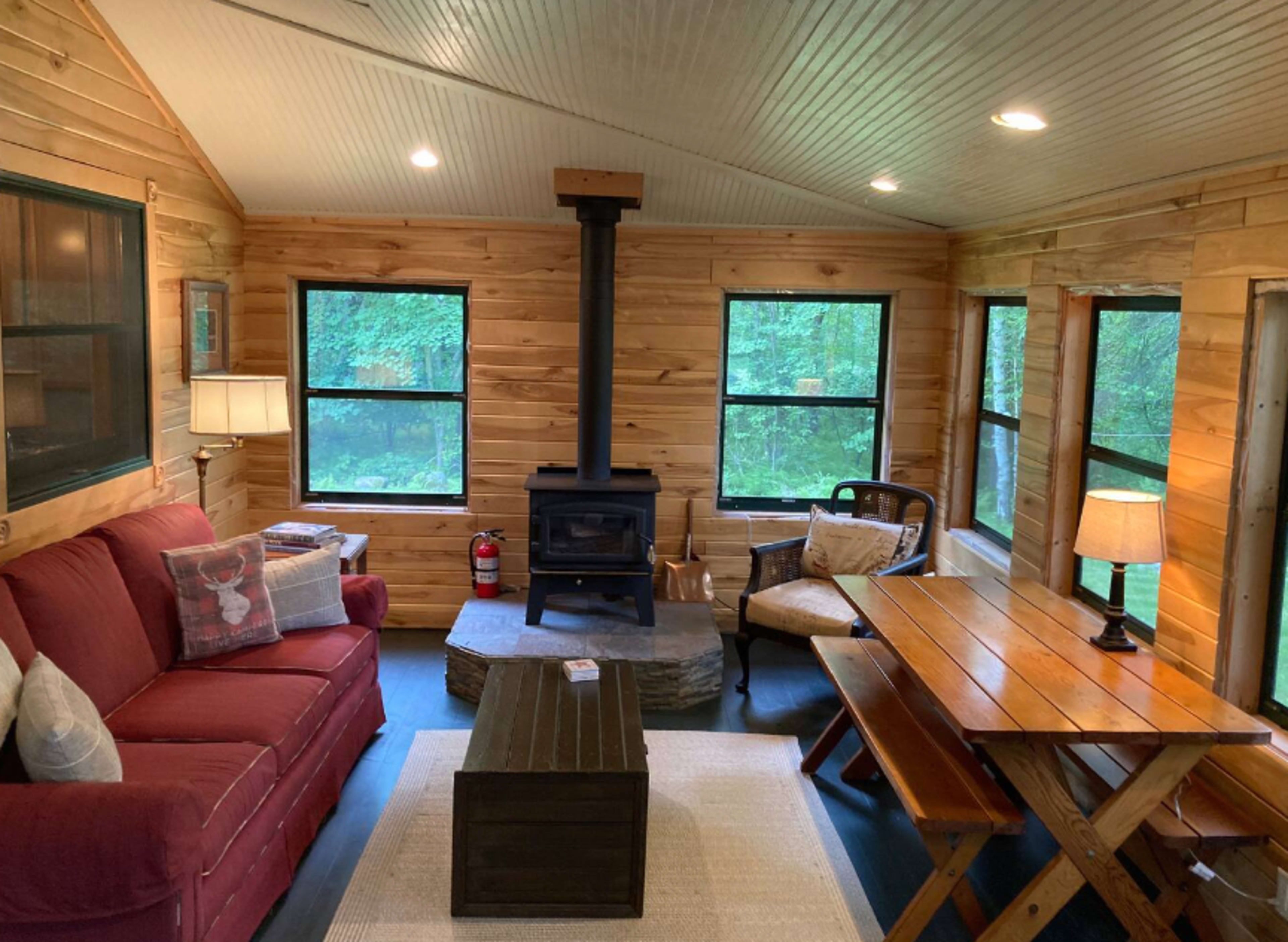 A cozy living area features a red sofa, a wood stove, a wooden dining table, and large windows overlooking a forested view.