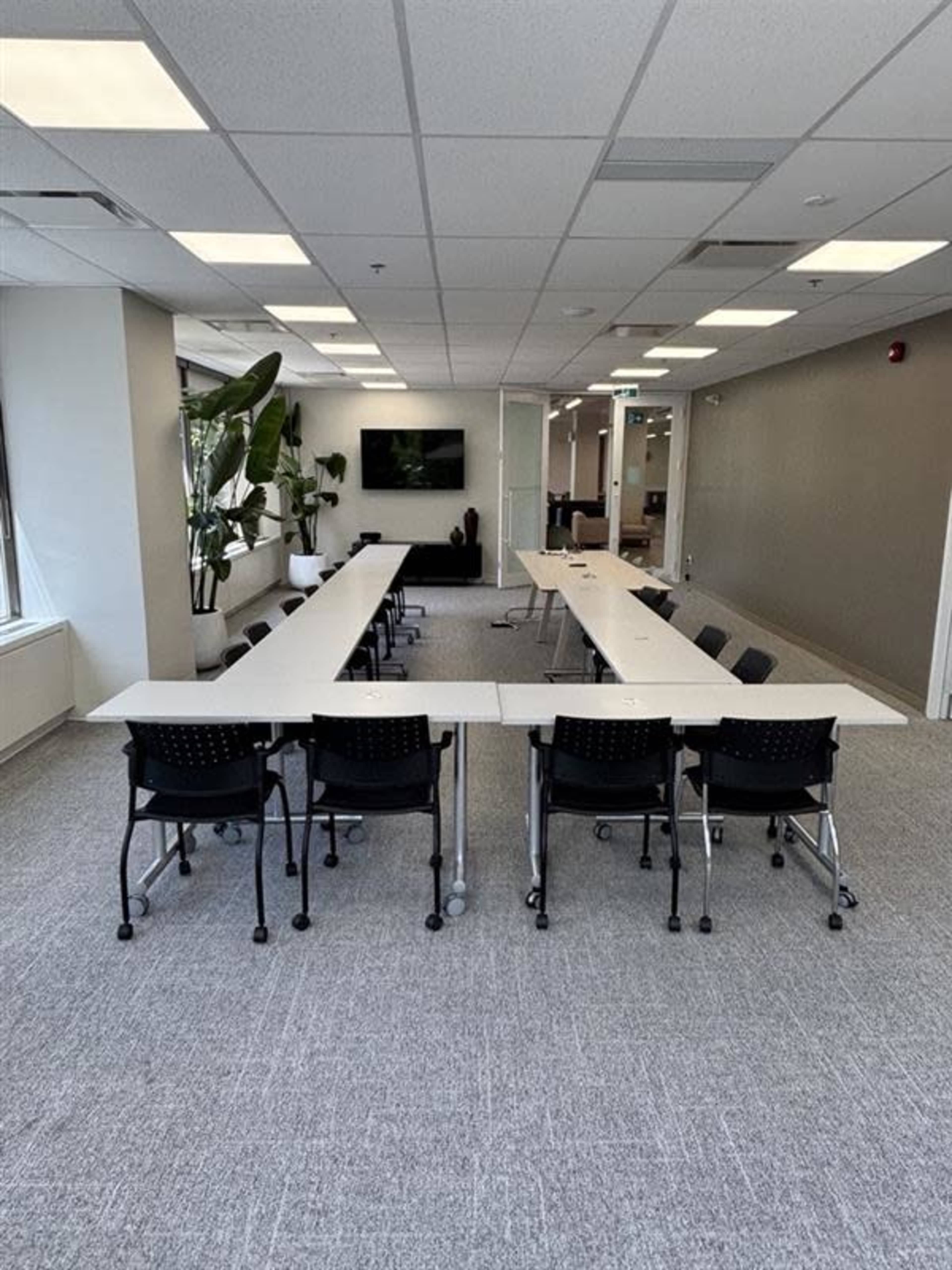 The image shows a modern conference room with several long tables arranged in a U-shape, accompanied by black rolling chairs and large windows.