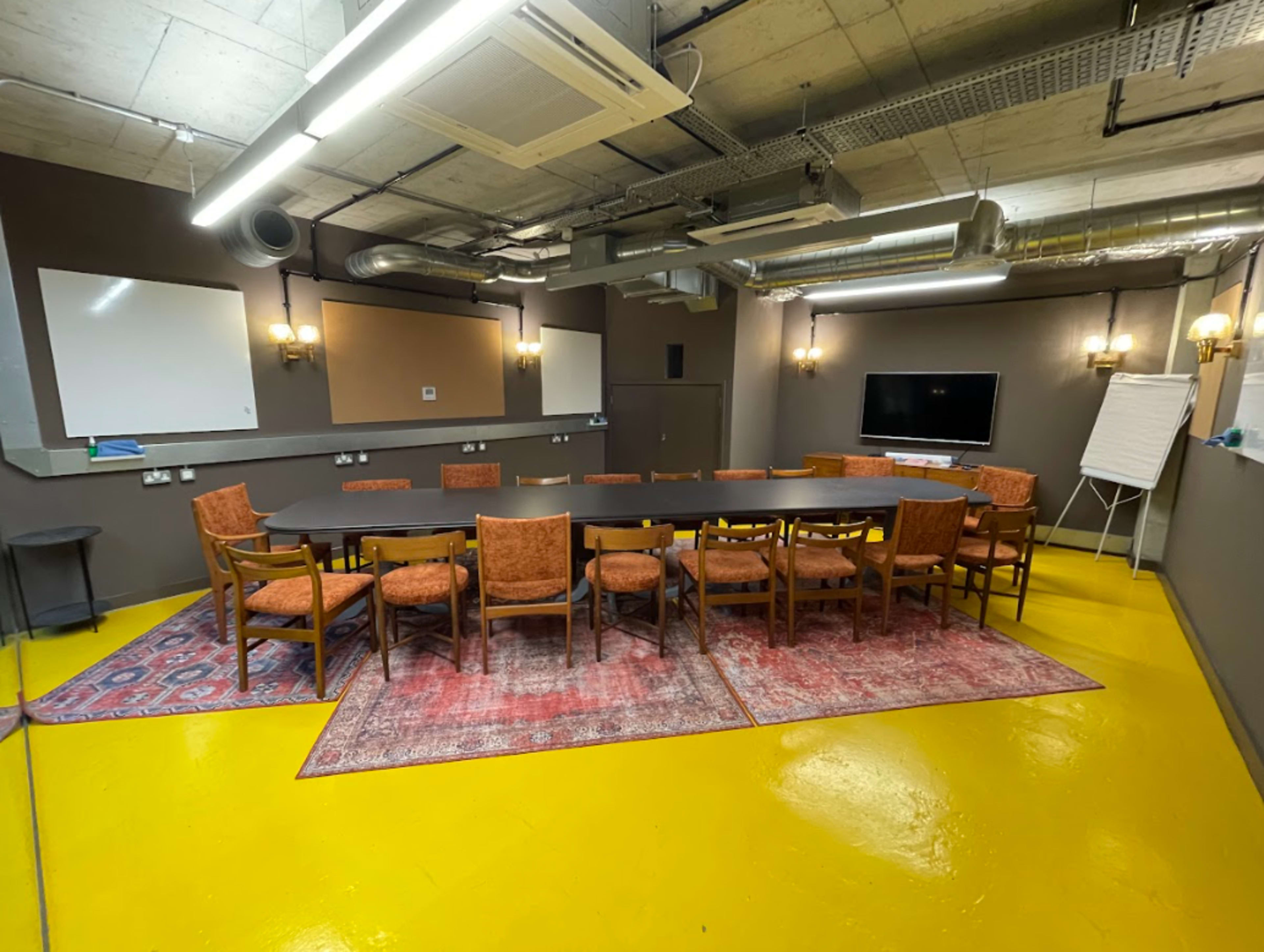 A rectangular conference room features a long table surrounded by chairs, with a large screen and whiteboards on the walls.