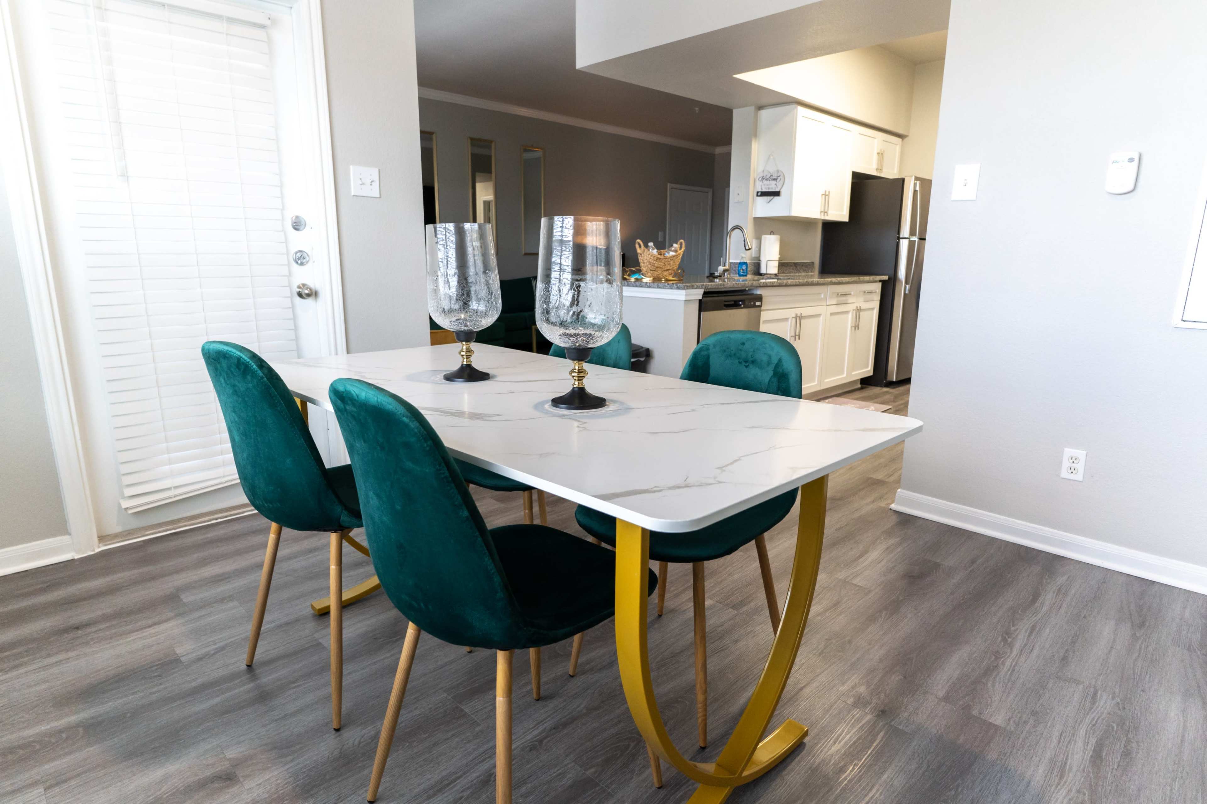 A modern dining area features a marble-topped table with gold legs surrounded by green upholstered chairs and two decorative glass candle holders.