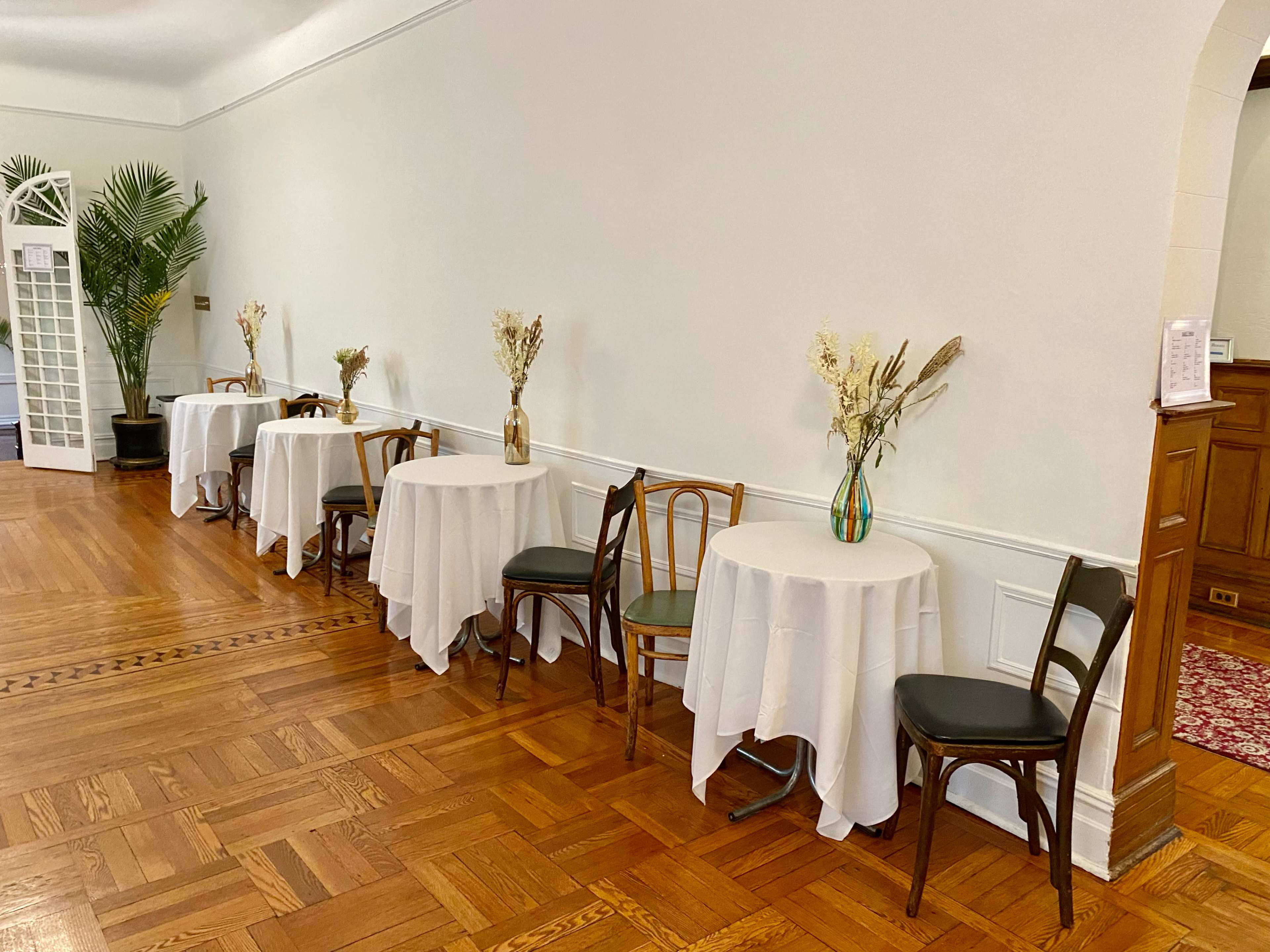 A row of tables covered with white tablecloths and adorned with vases of dried flowers is lined against a wall in a room with wooden flooring.