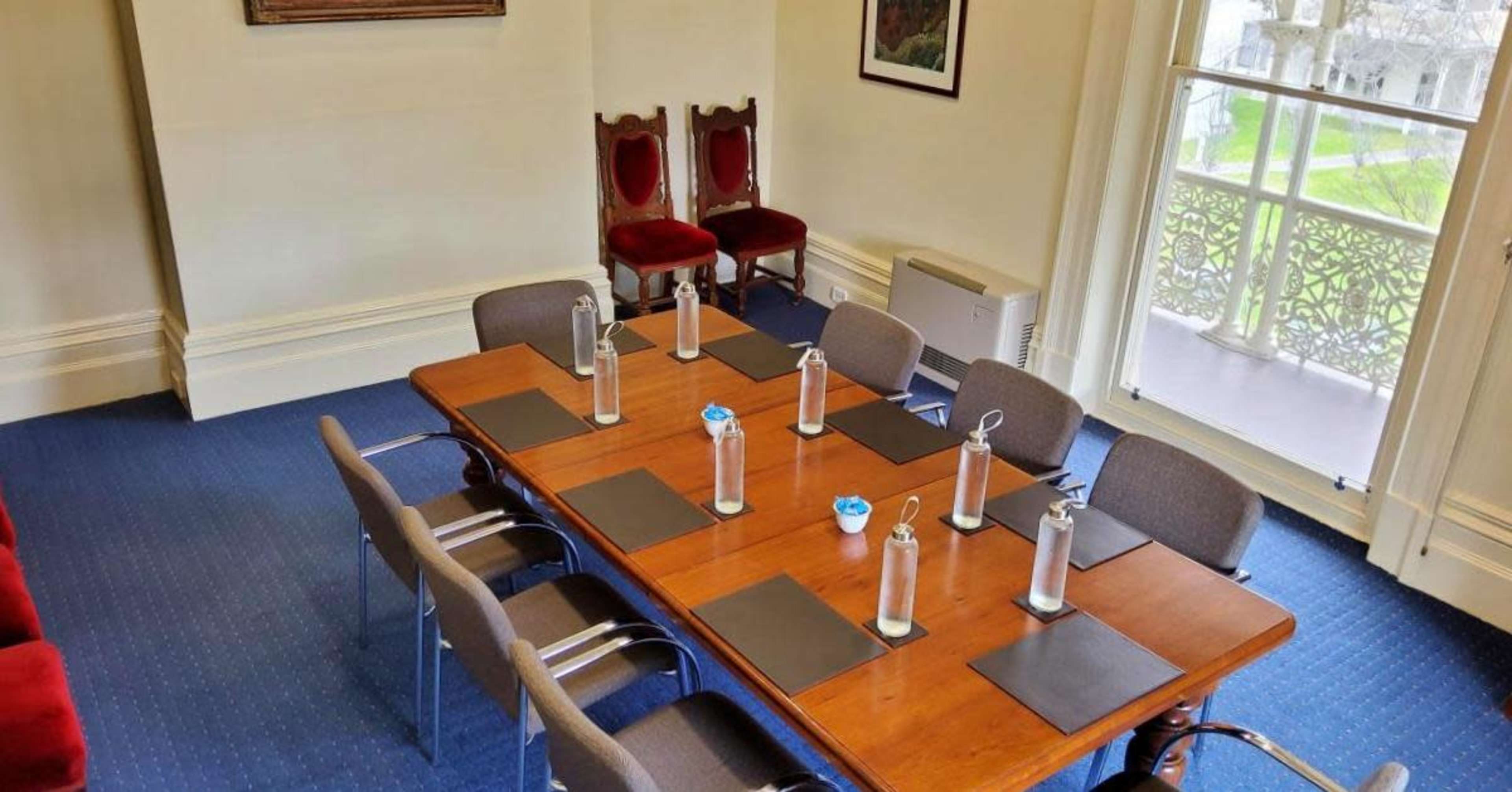 A conference room features a wooden table with water bottles and place mats, surrounded by chairs and two red upholstered chairs against the wall.