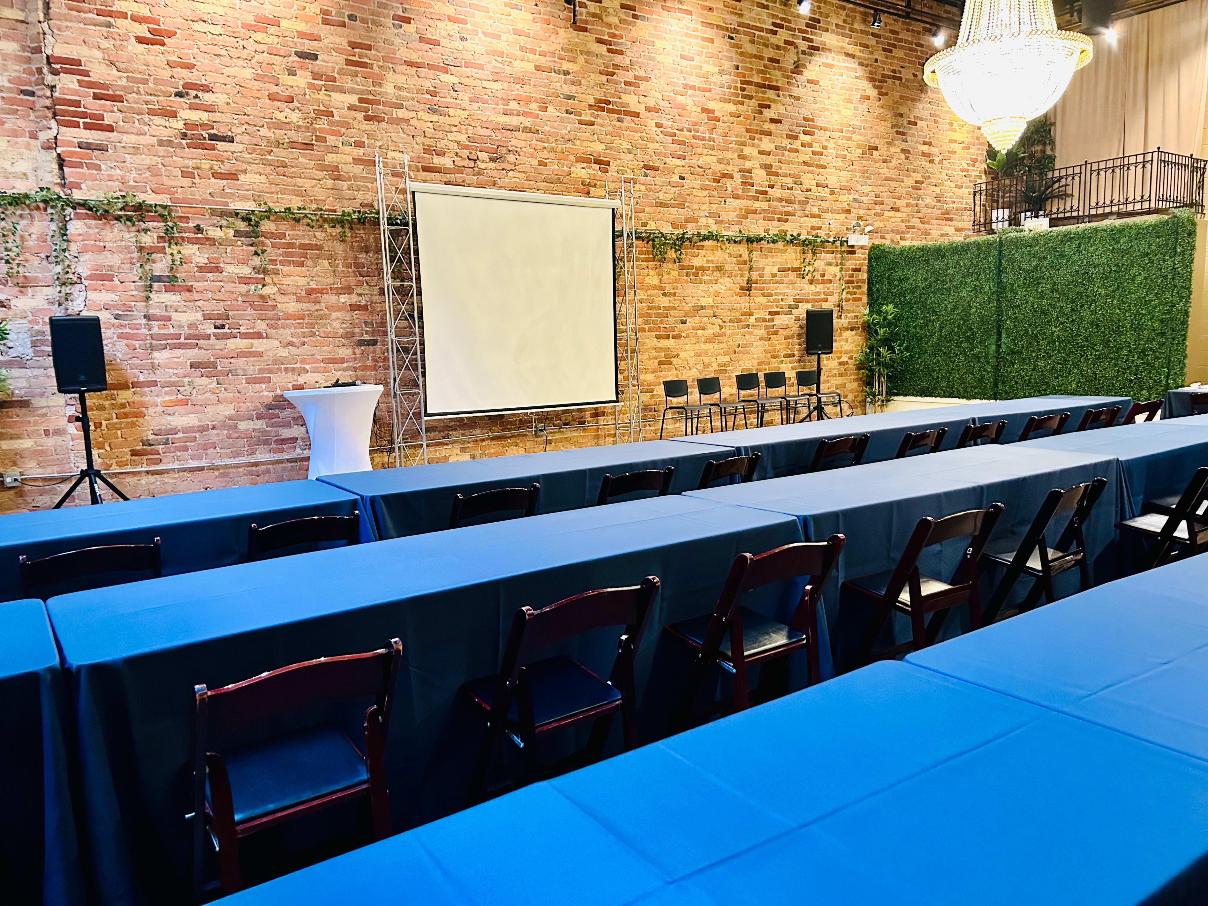 The image shows a conference room with blue tables arranged in rows, a blank presentation screen on a brick wall, and greenery in the background.