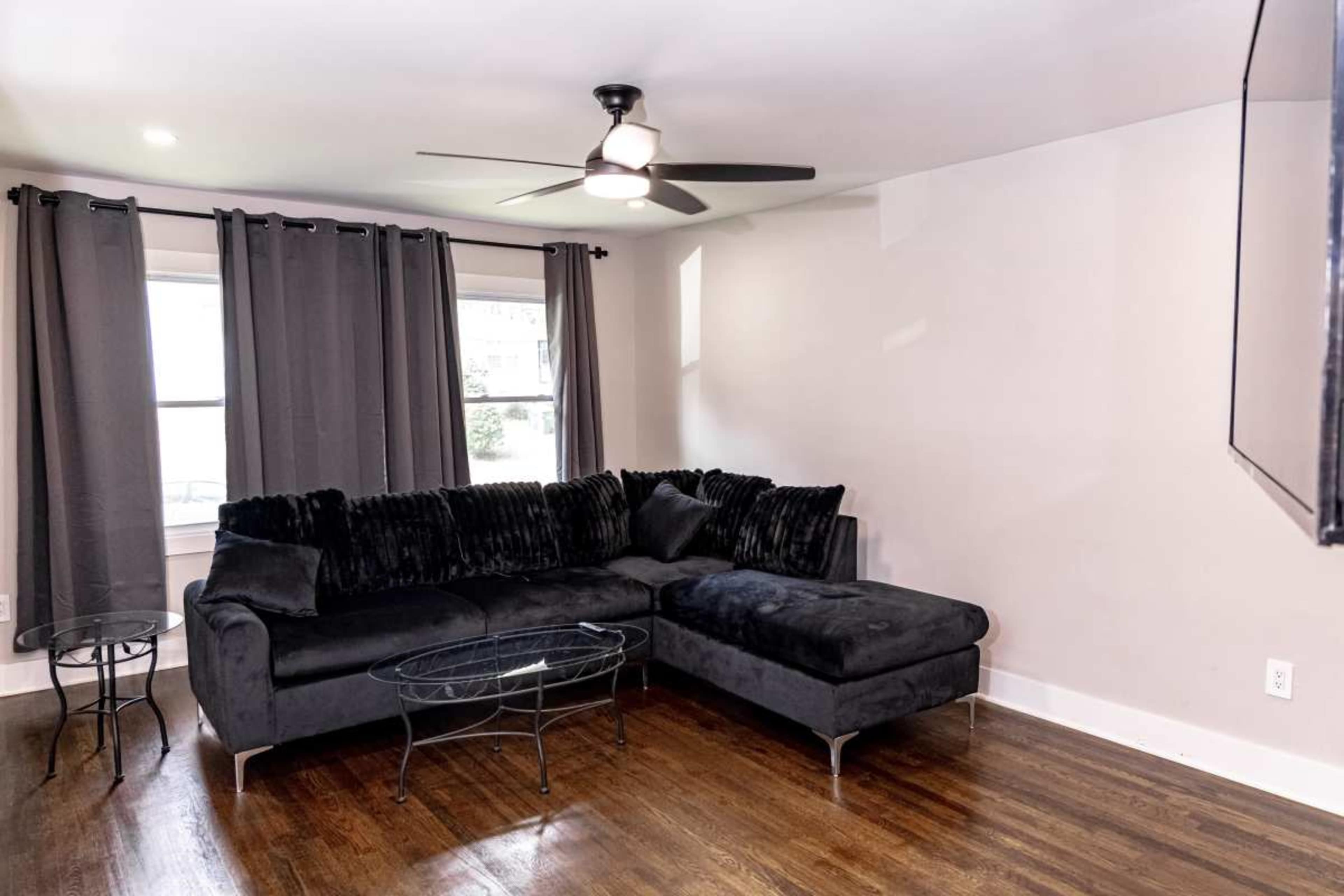The image shows a modern living room featuring a black sectional sofa, a glass side table, and gray curtains over a window.