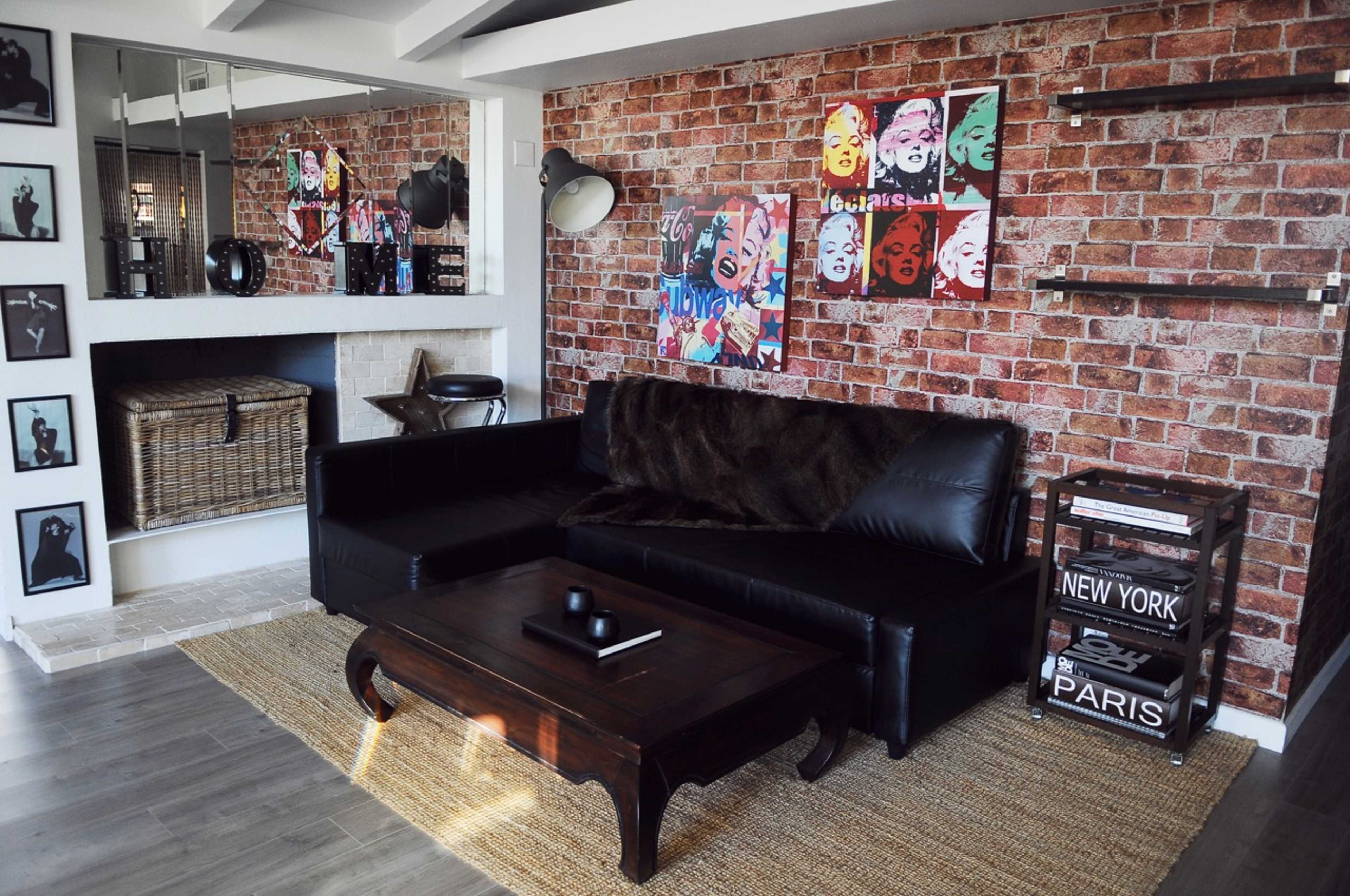 A modern living room features a black leather sectional sofa, a dark wooden coffee table, and a brick wall adorned with colorful artwork.