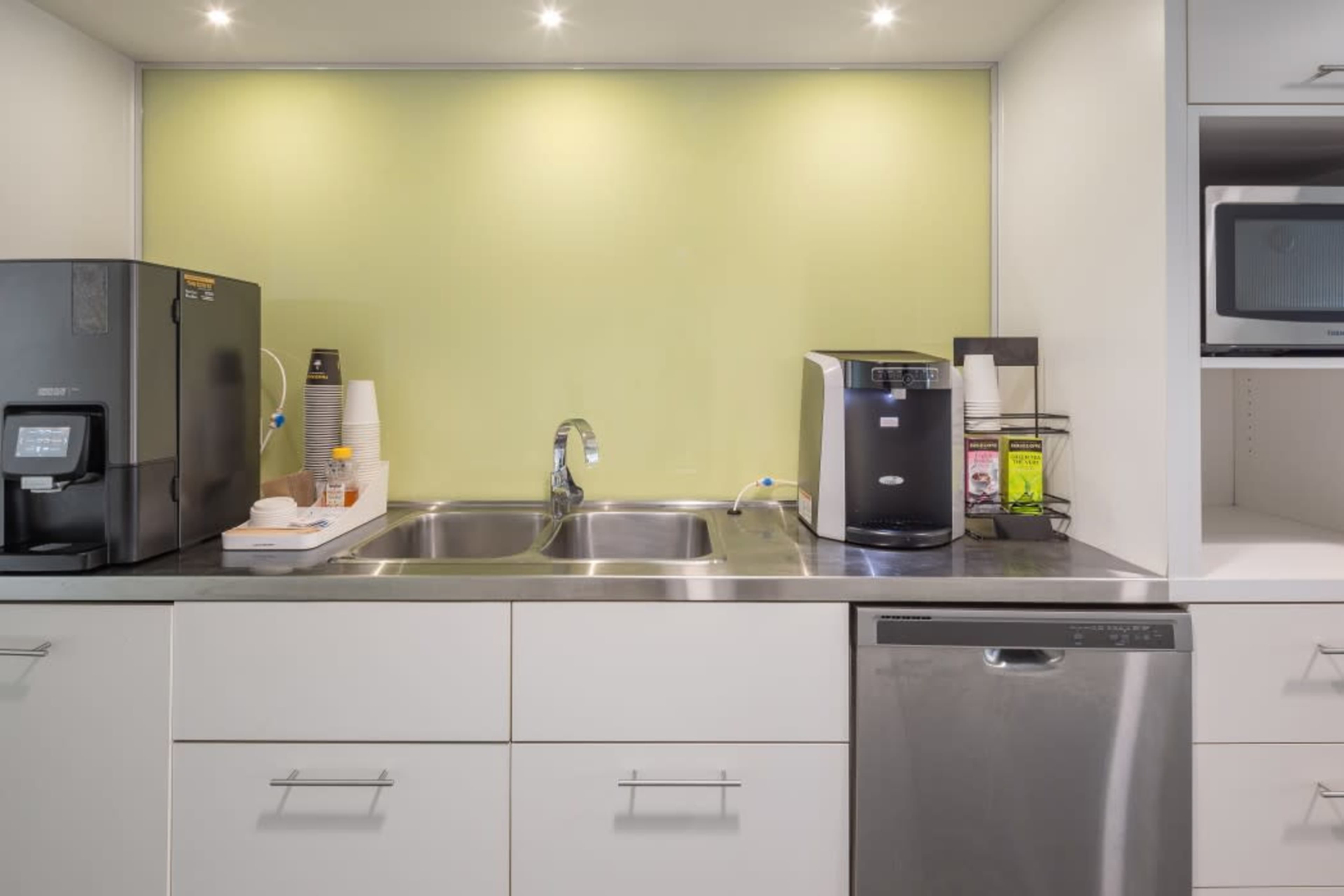 A modern kitchen area with a double sink, a coffee machine, and a dishwasher, set against a light green wall.
