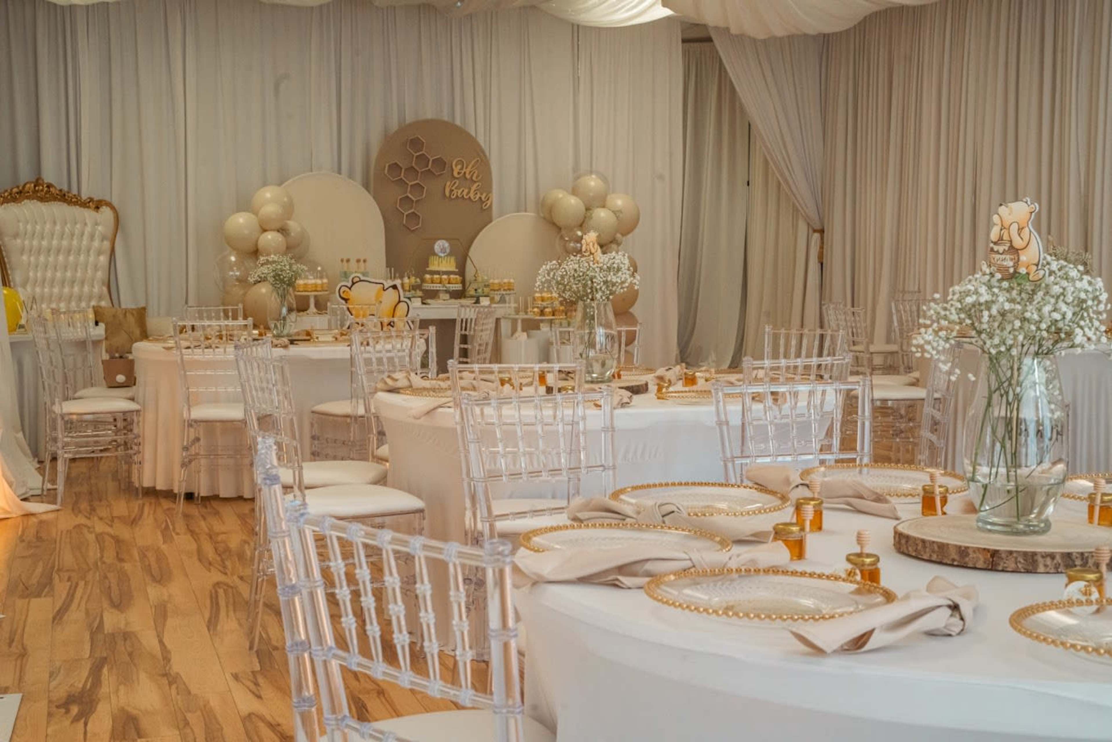 The image shows an elegantly decorated event space with round tables, white tablecloths, clear chiavari chairs, and decorative elements including floral arrangements and balloons.
