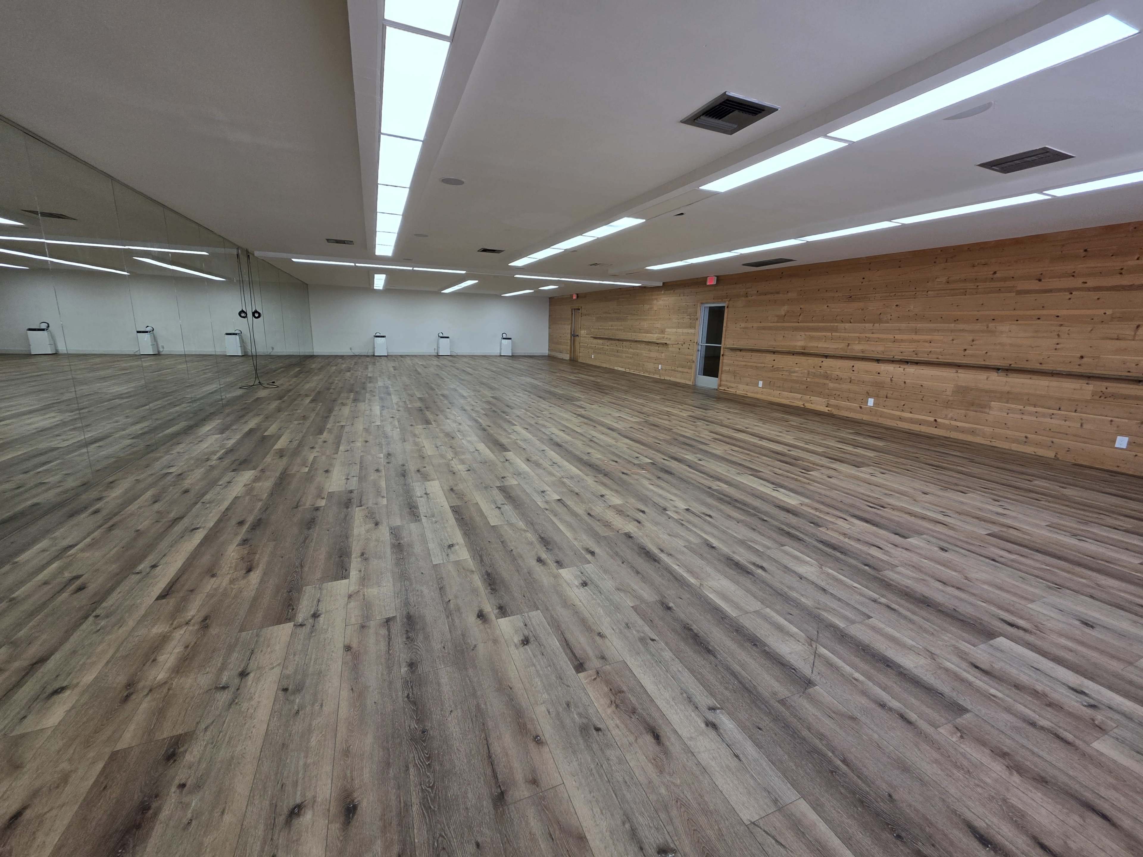 The image shows a spacious, empty room with wooden floors and one mirrored wall.