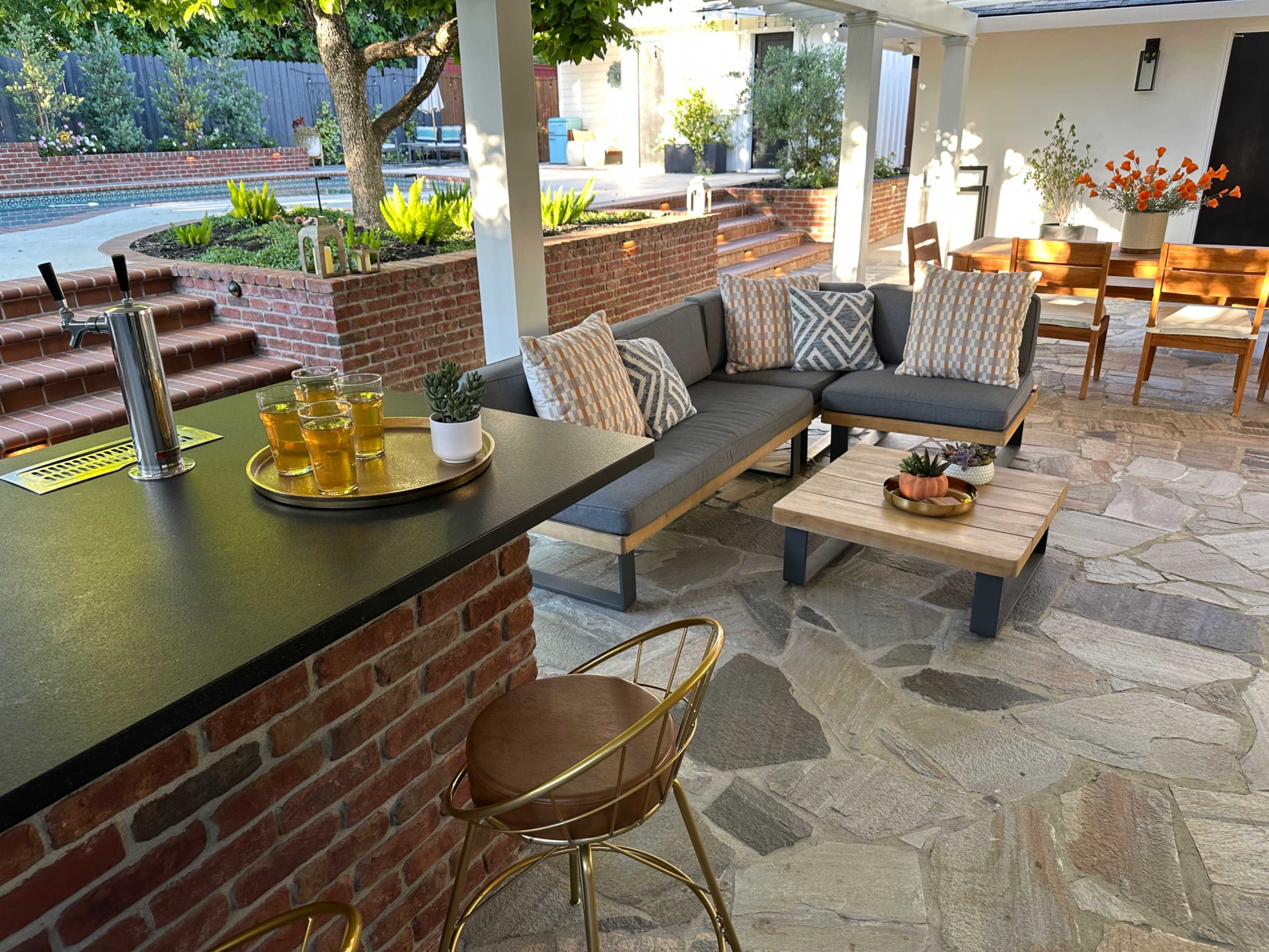 The image shows a modern outdoor patio area featuring a comfortable seating arrangement, a bar with drinks, and a landscaped background with steps leading to a swimming pool.