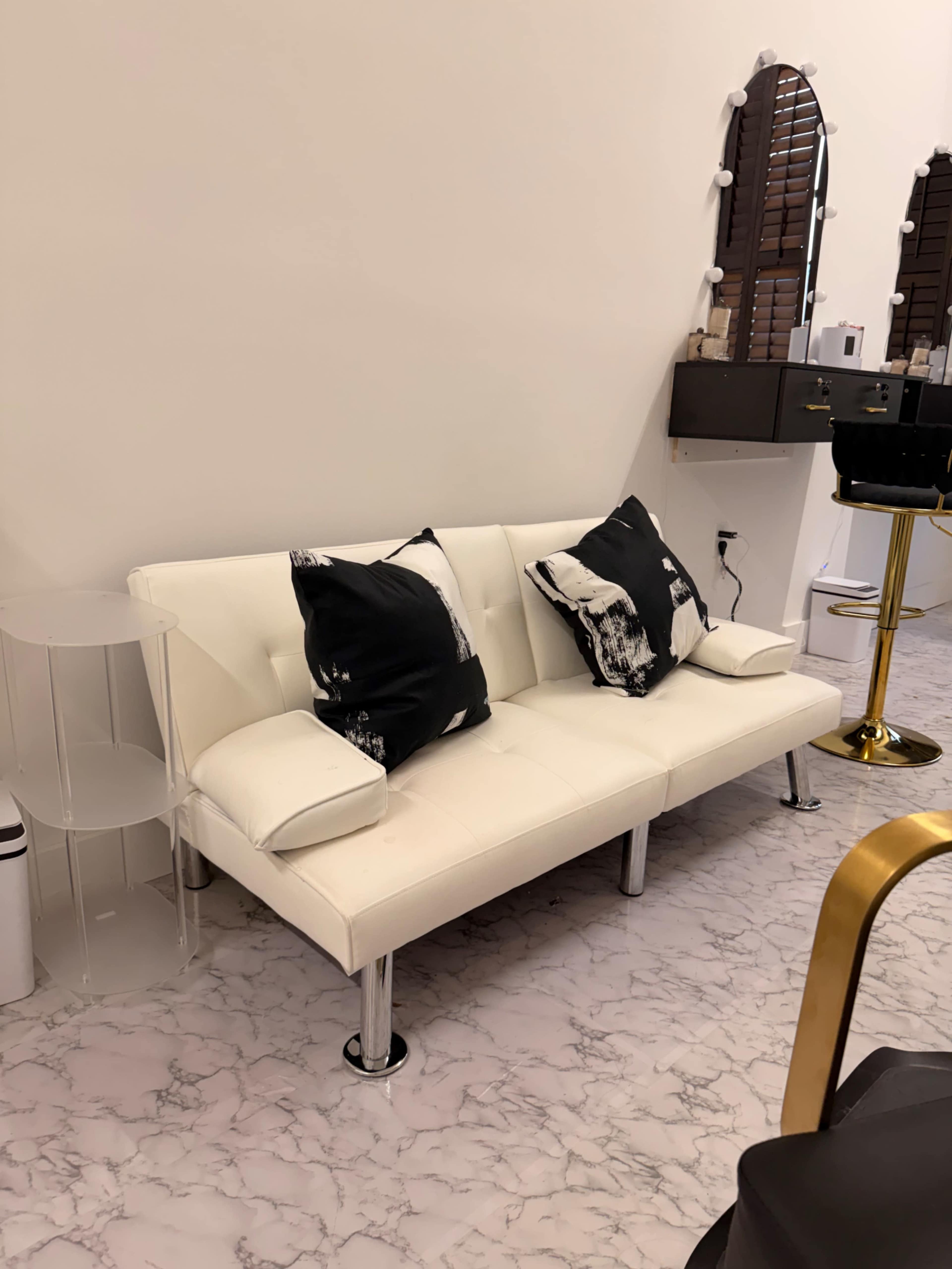 The image shows a white sofa with black and white patterned pillows placed against a light-colored wall, accompanied by a clear side table and a stylized mirror setup.