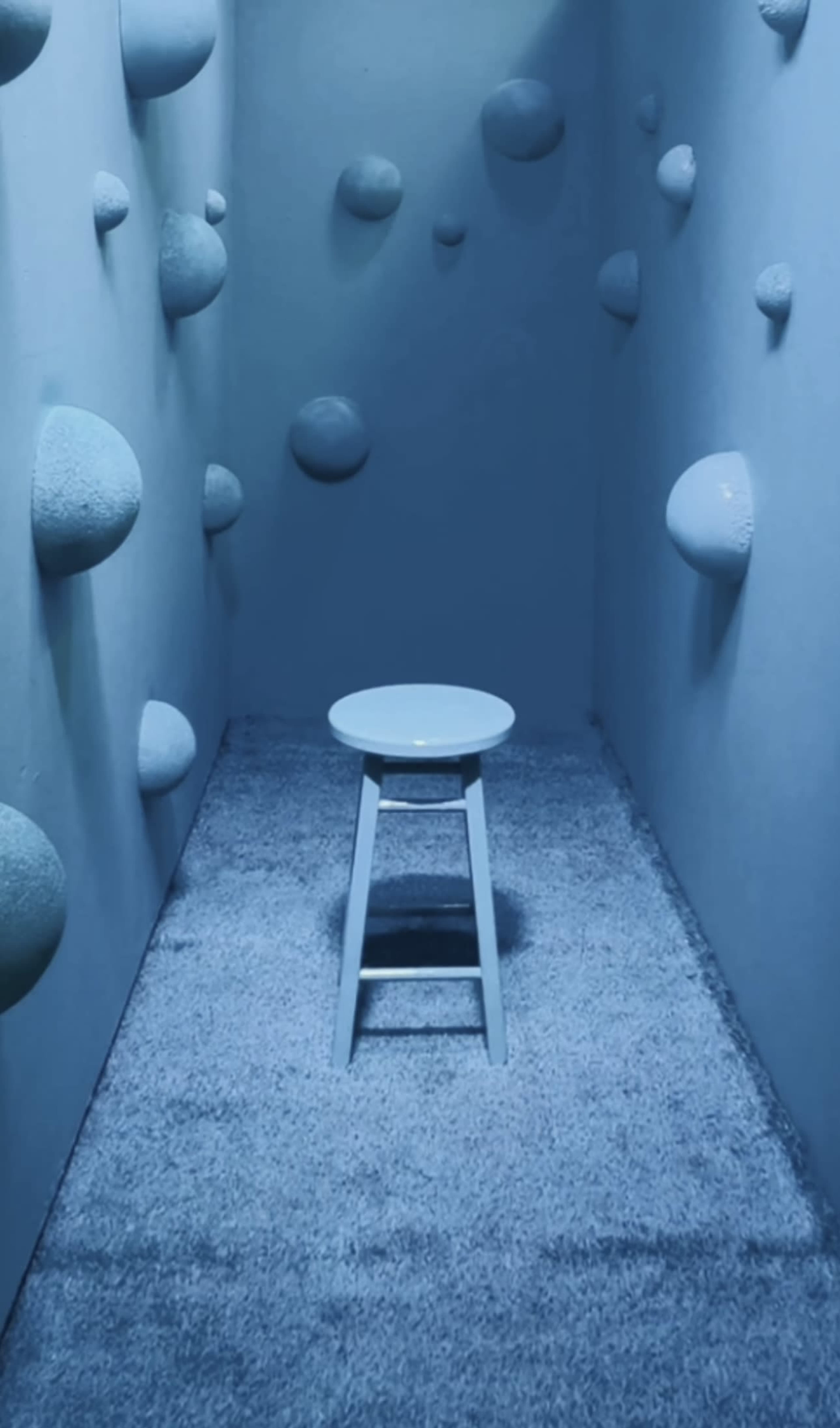 A blue-walled room features a single stool in the center, surrounded by spherical objects on the walls.