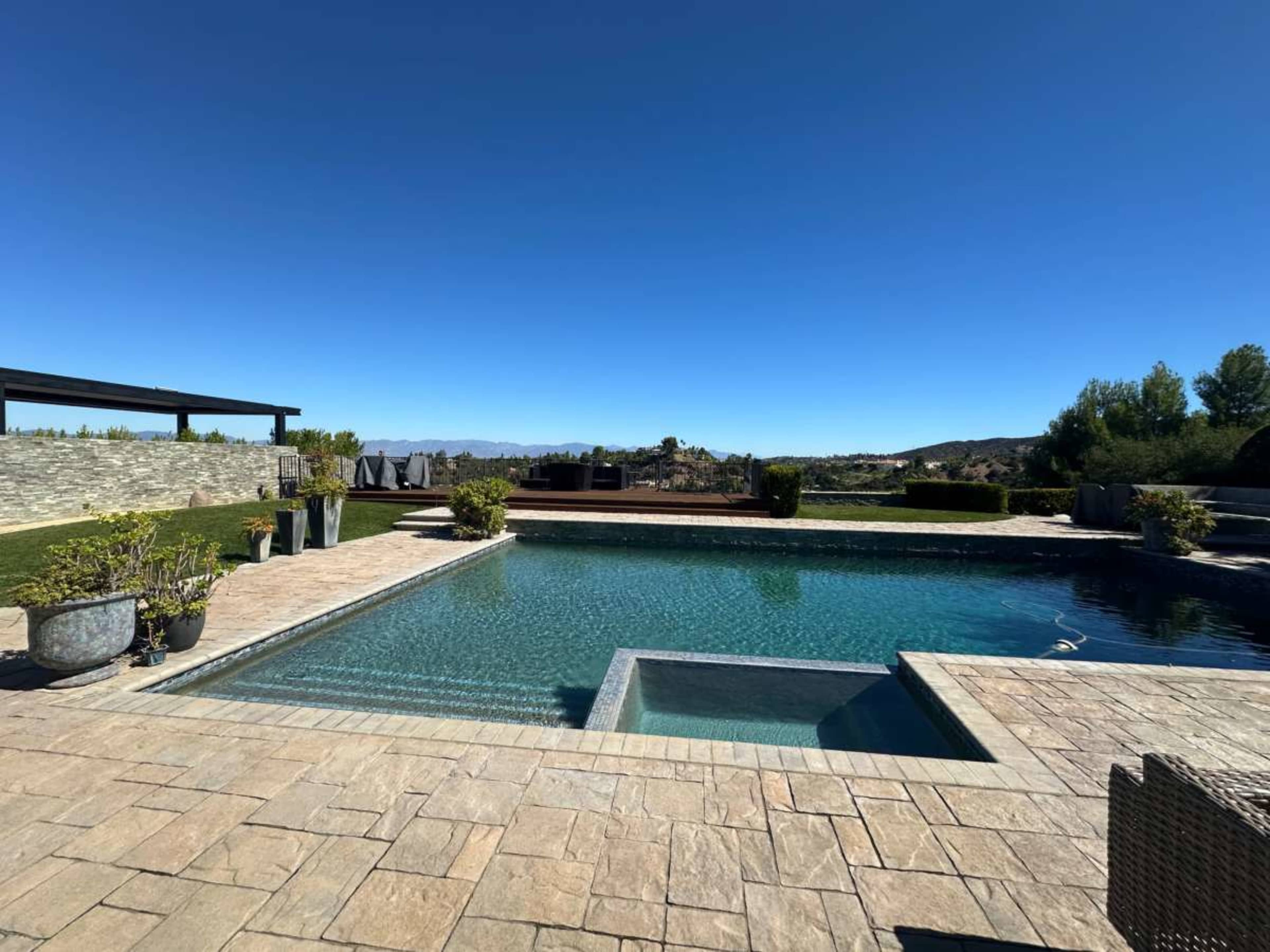 The image shows a clear swimming pool surrounded by stone patios and landscaped grounds under a blue sky.