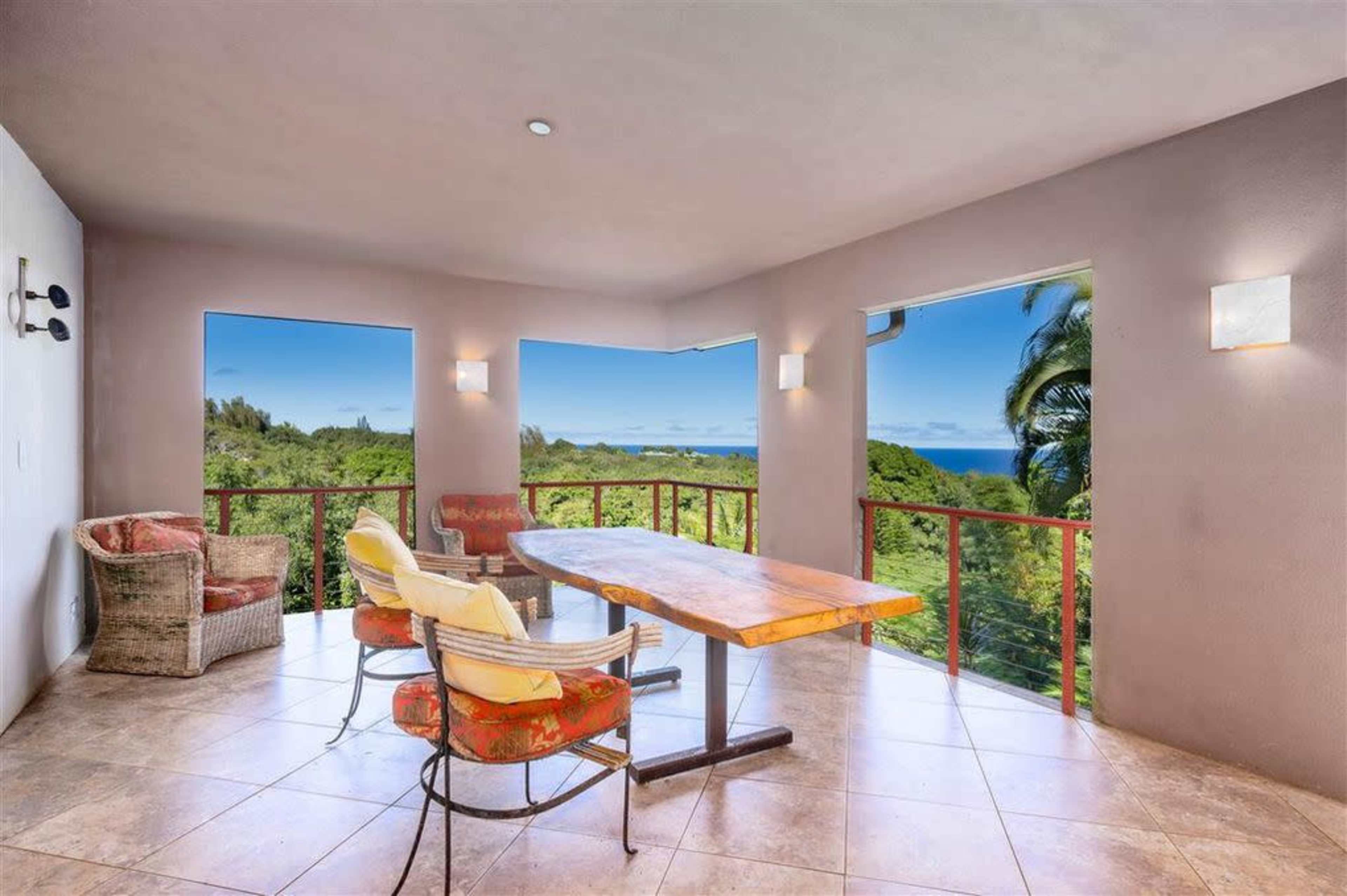 A spacious, sunlit room features a large wooden table surrounded by chairs, with panoramic windows showcasing a view of greenery and the ocean.