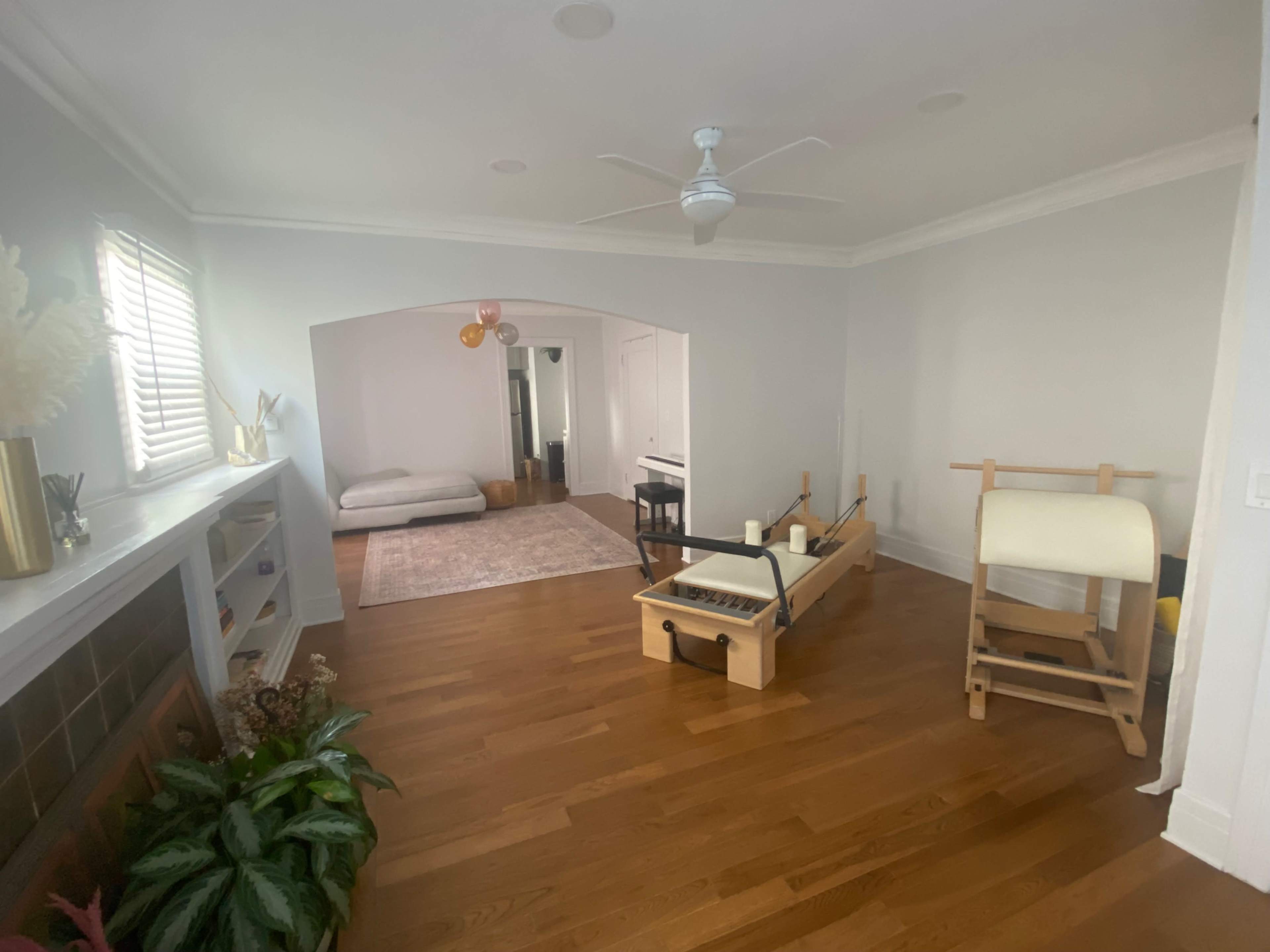The image shows a spacious room with wooden flooring, featuring a reformer Pilates machine, a small area rug, and a gray sofa in the background.