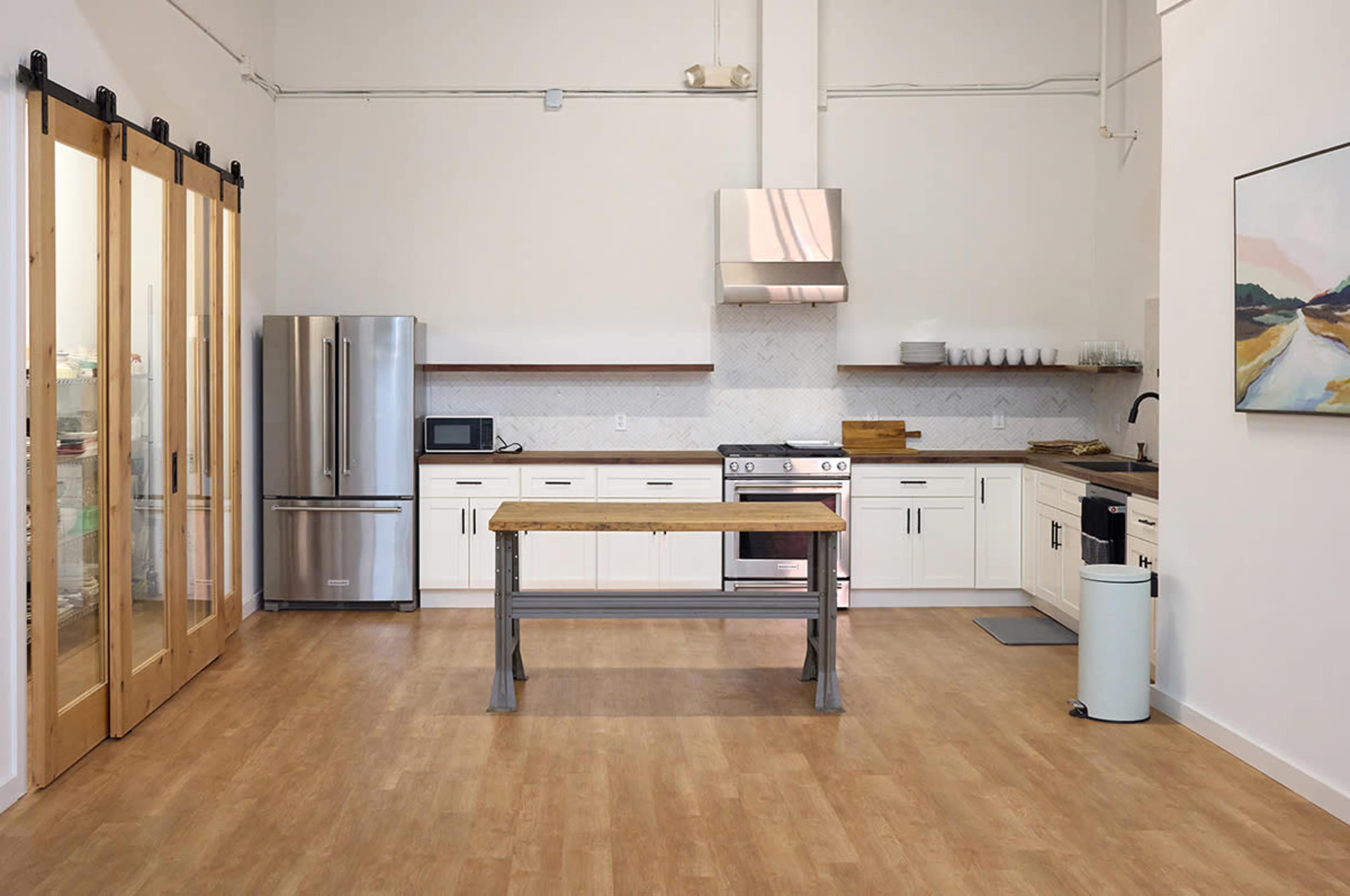 The kitchen features stainless steel appliances, a central wooden table, and white cabinetry with a light wood floor.