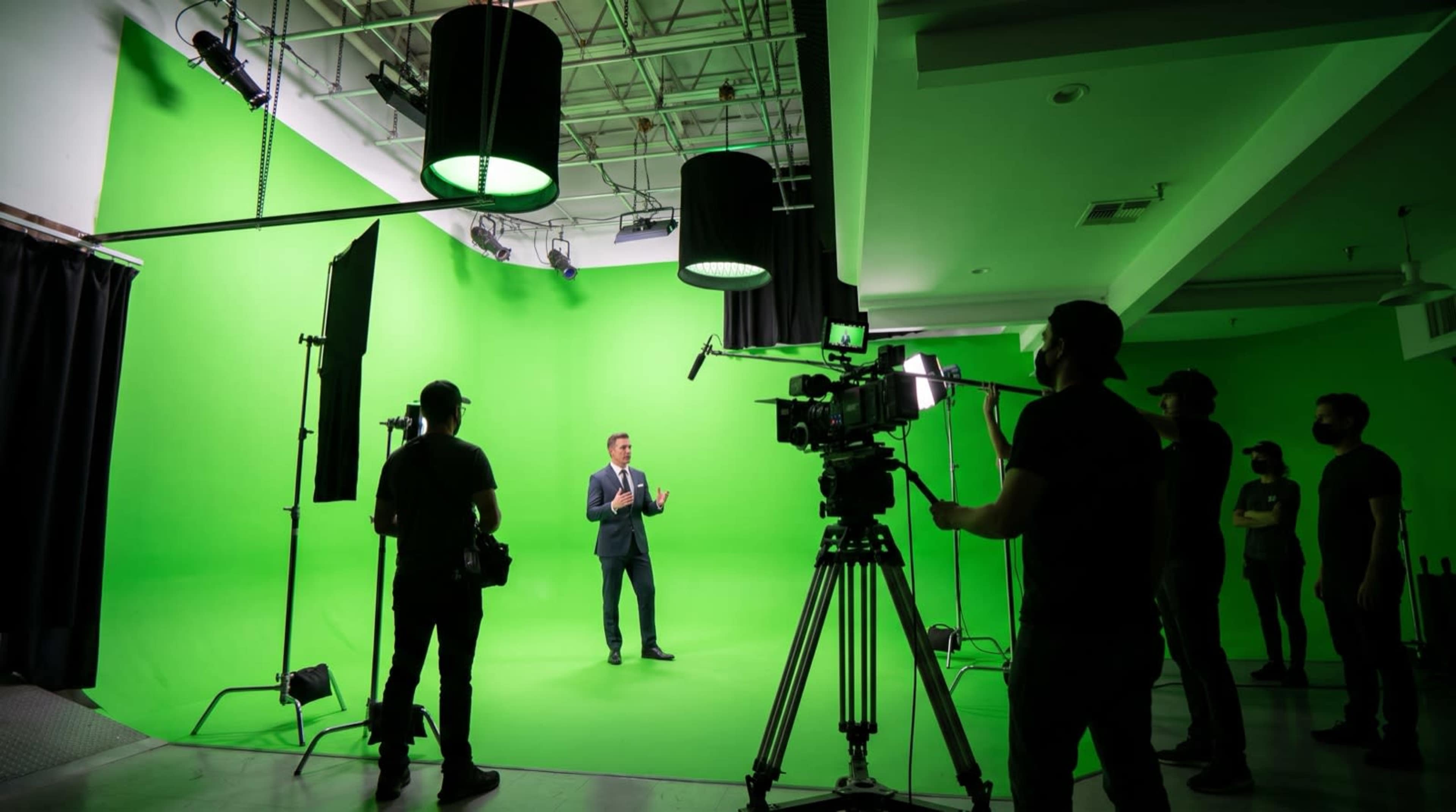 A filming crew sets up equipment around a man in a suit standing in front of a green screen in a studio.