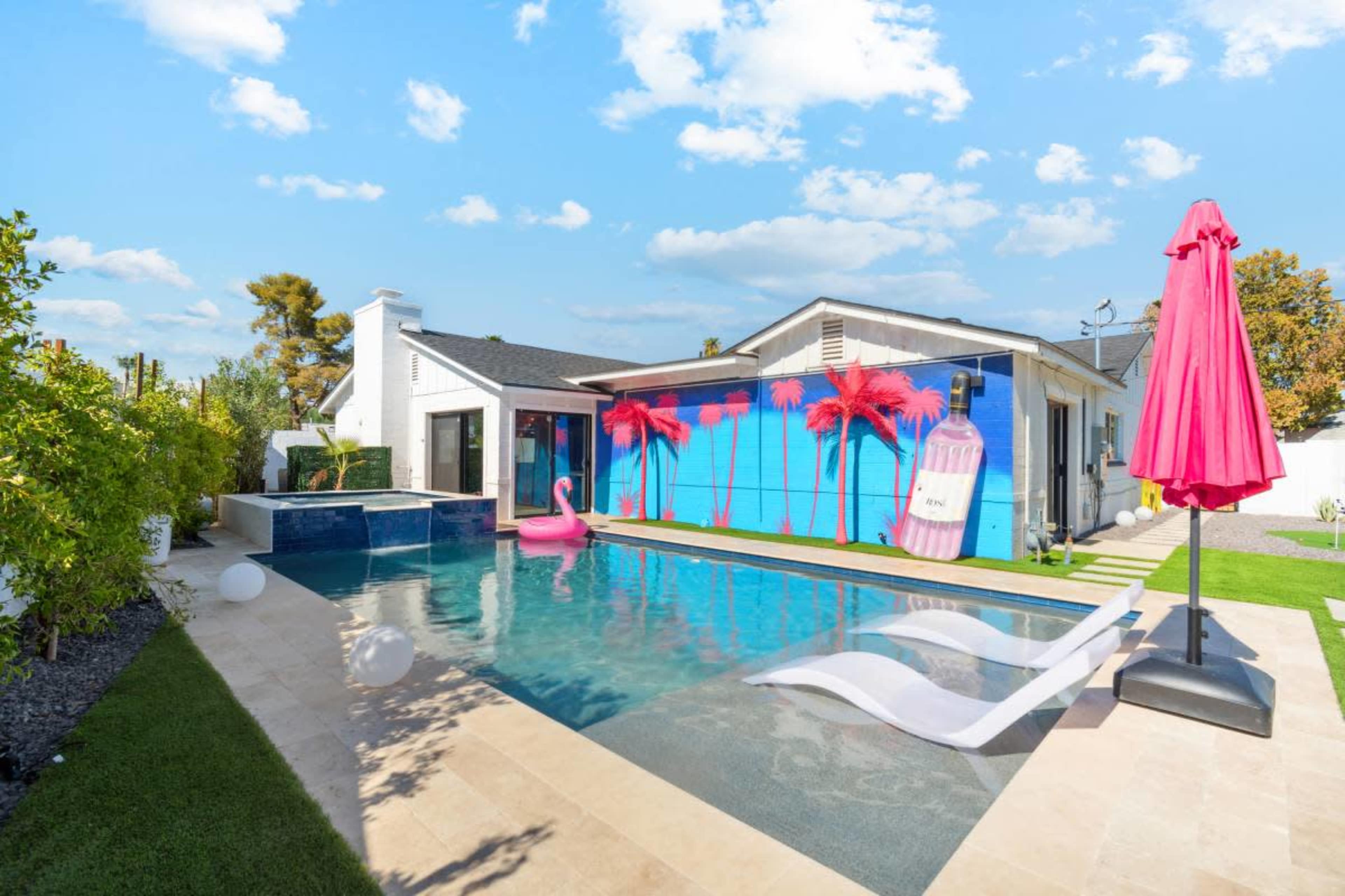 The image shows a modern backyard with a swimming pool, palm tree murals on a wall, a pink flamingo float, and a large pink umbrella beside lounge chairs.