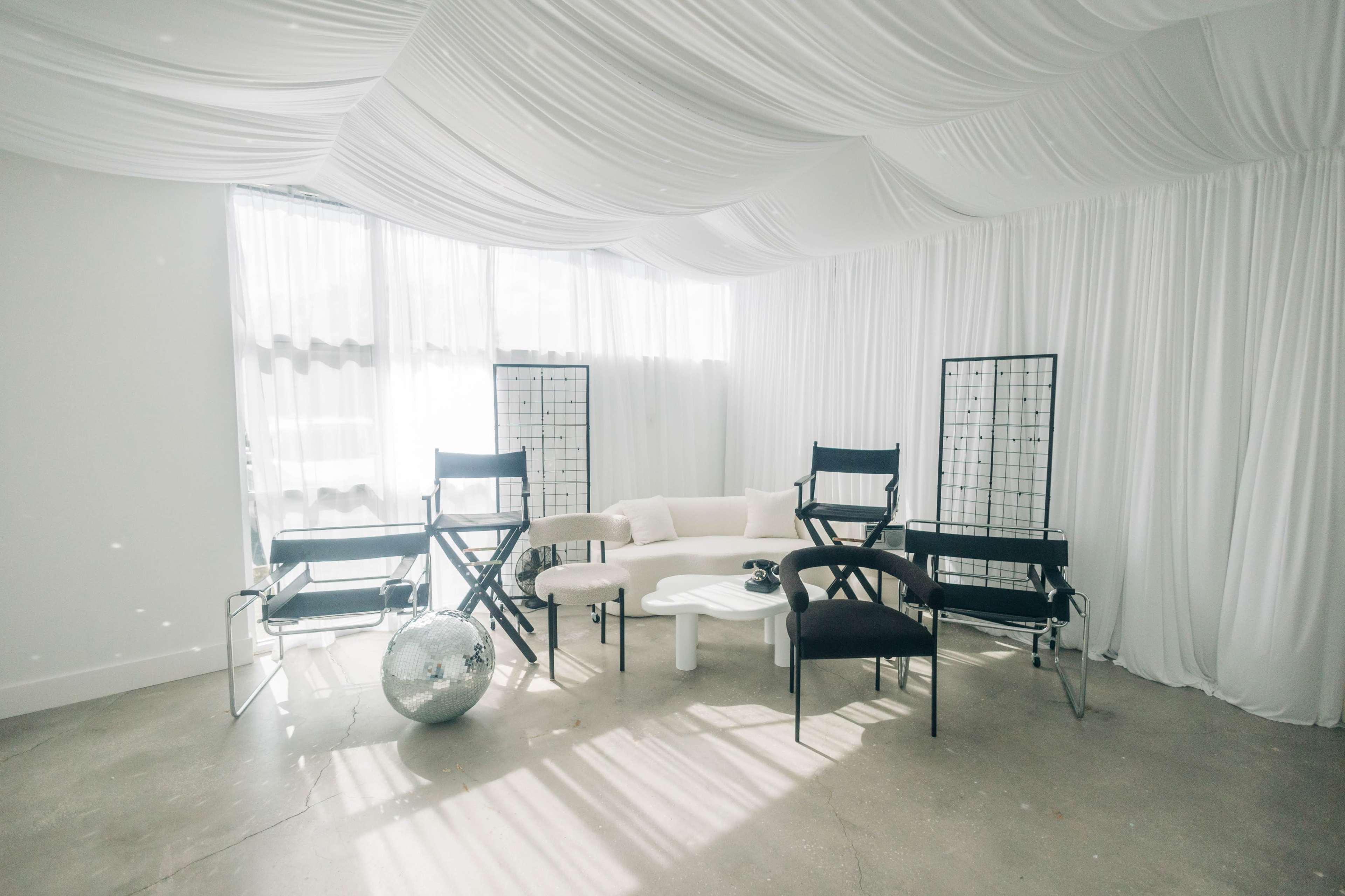 The image shows a modern, minimalist room featuring black director's chairs and a white circular sofa surrounded by soft white drapery and a mirrored disco ball.