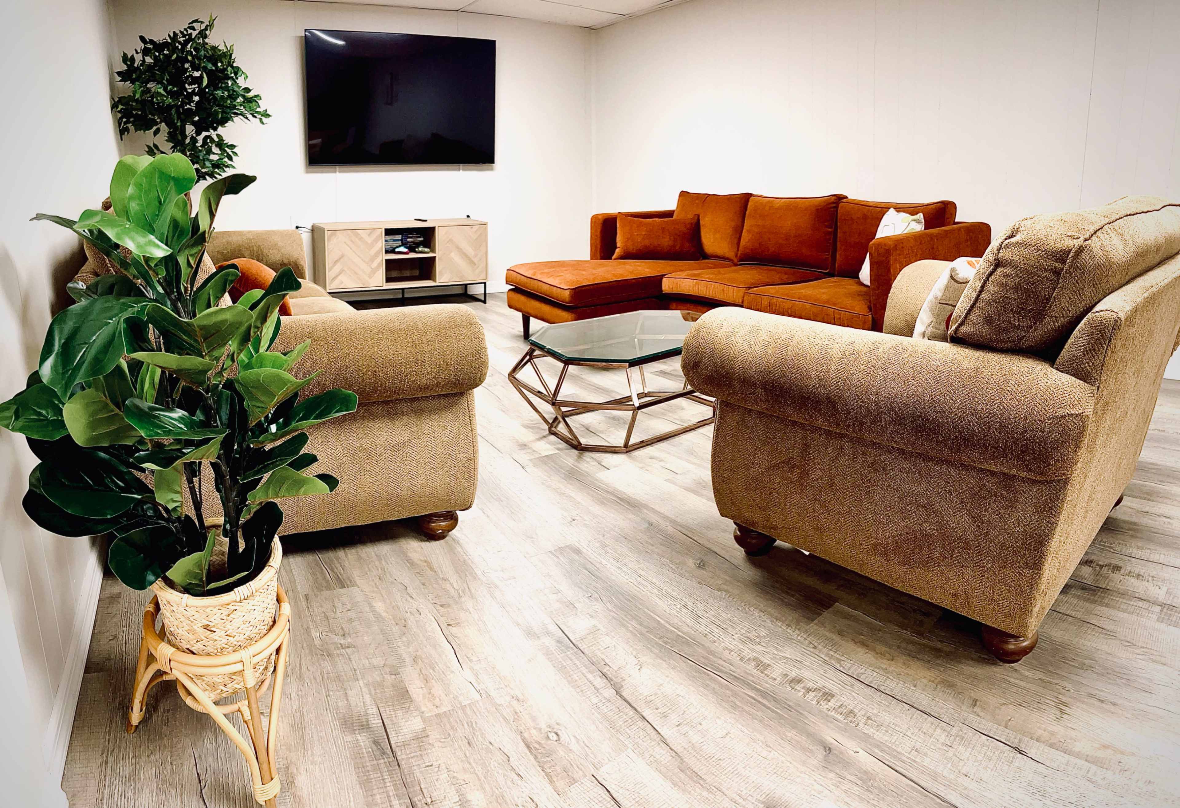 A modern, low-lit living room features a large TV mounted on the wall, a glass coffee table, and an arrangement of orange and brown sofas with a decorative plant in a wicker pot.
