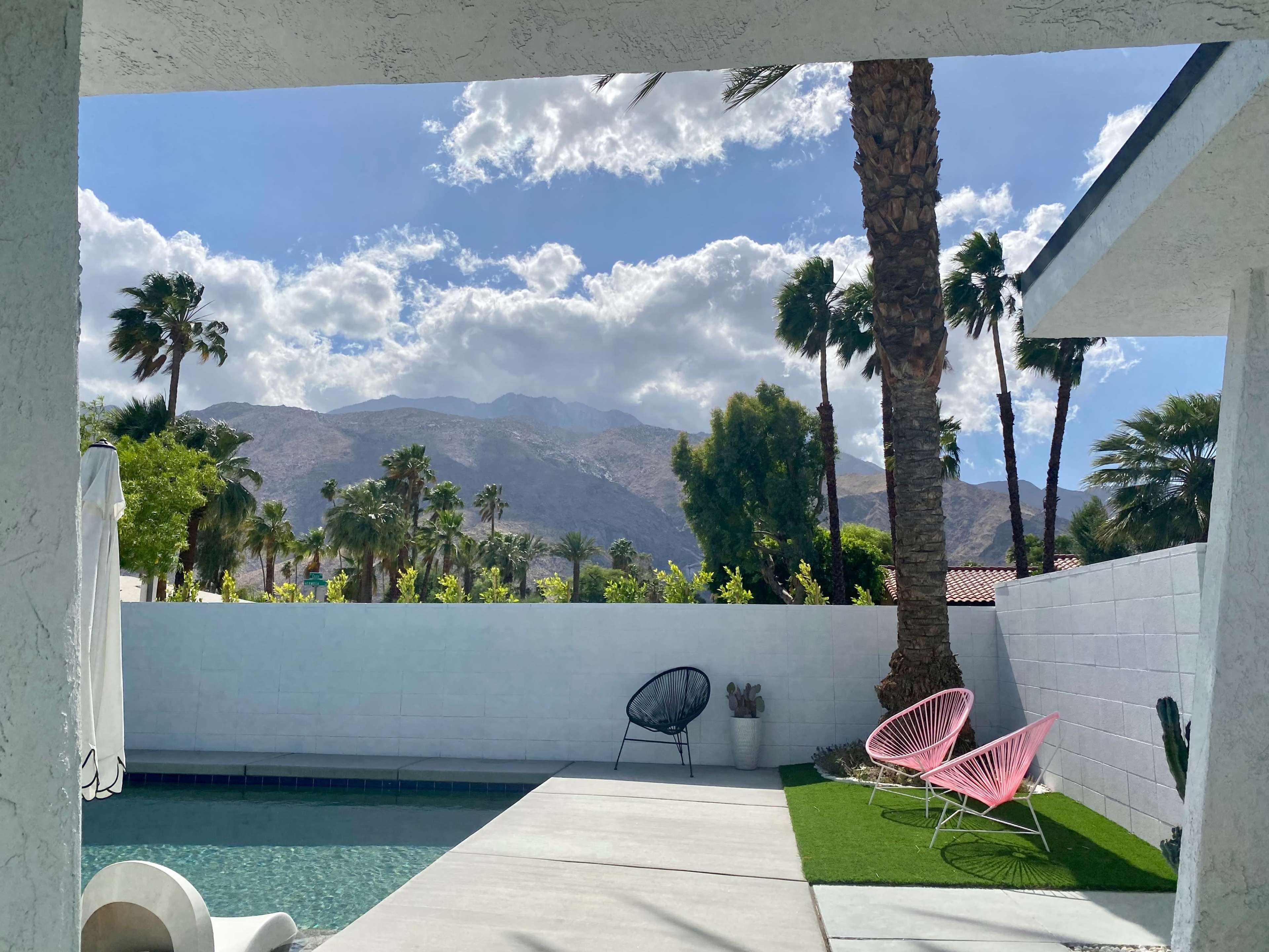 A poolside view with colorful chairs set against a backdrop of palm trees and mountains under a partly cloudy sky.