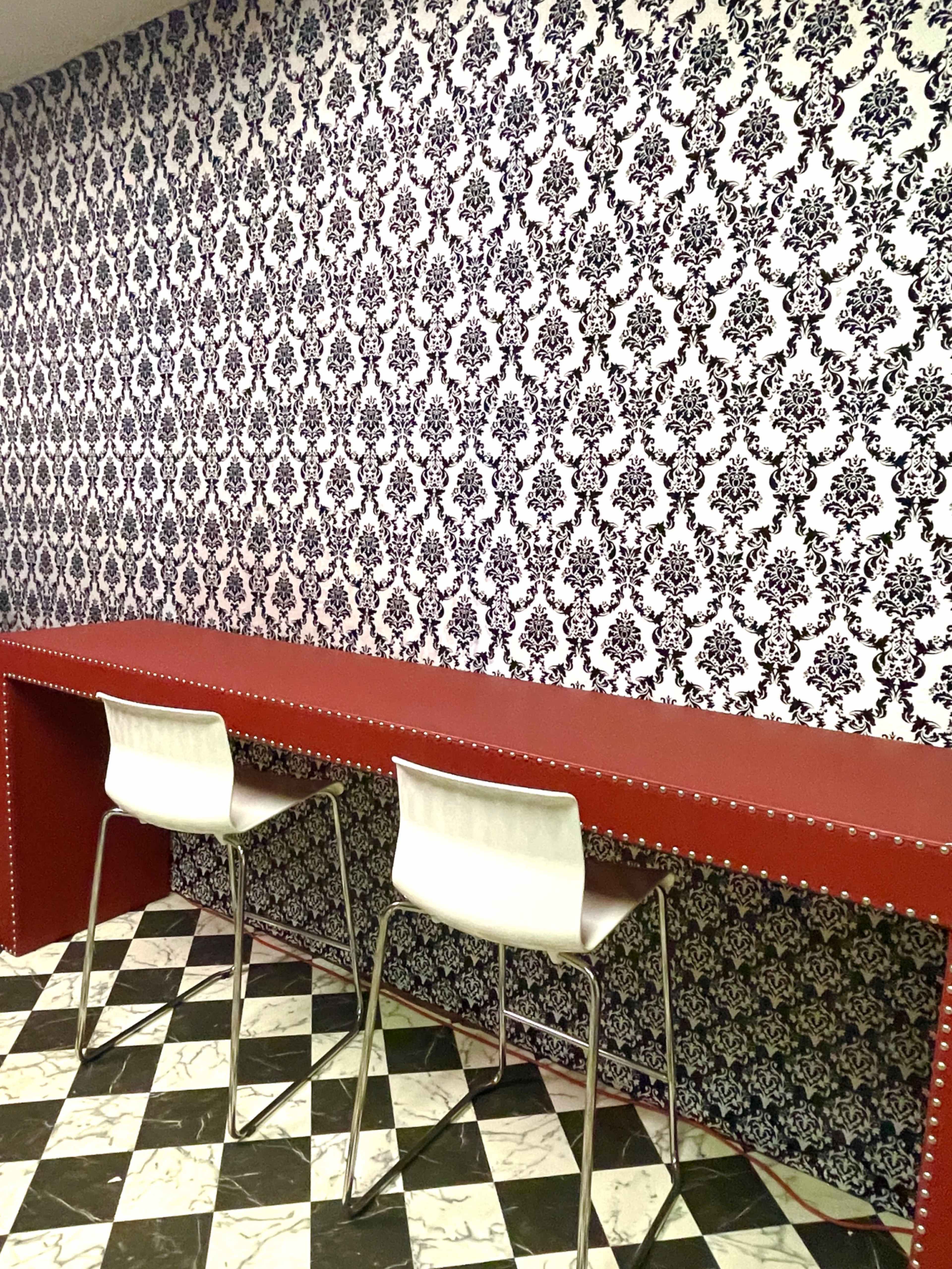 A room with a patterned black and white wallpaper, a red counter, and two white chairs against a tiled floor of alternating black and white diamond shapes.