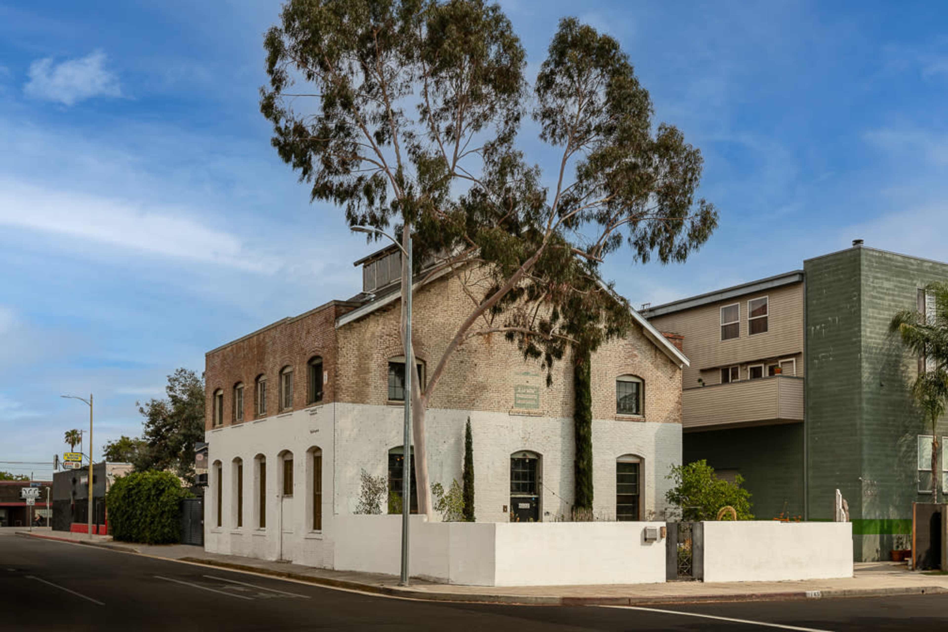 A two-story brick building with large windows is situated at the corner of a street, adjacent to a modern green structure and a tall tree.