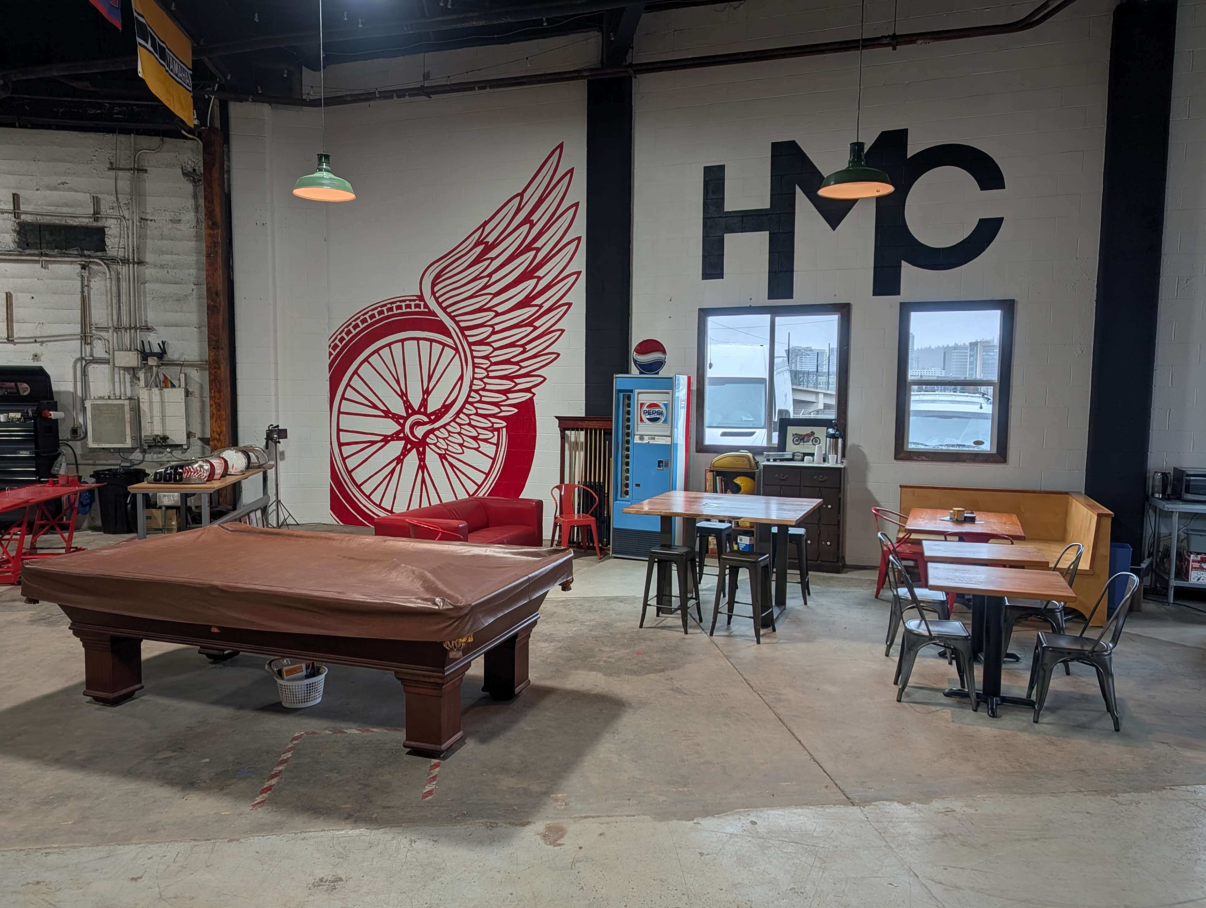 The image shows a spacious industrial room with a pool table covered by a brown cloth, several tables and chairs, and a large winged bicycle mural on the wall.