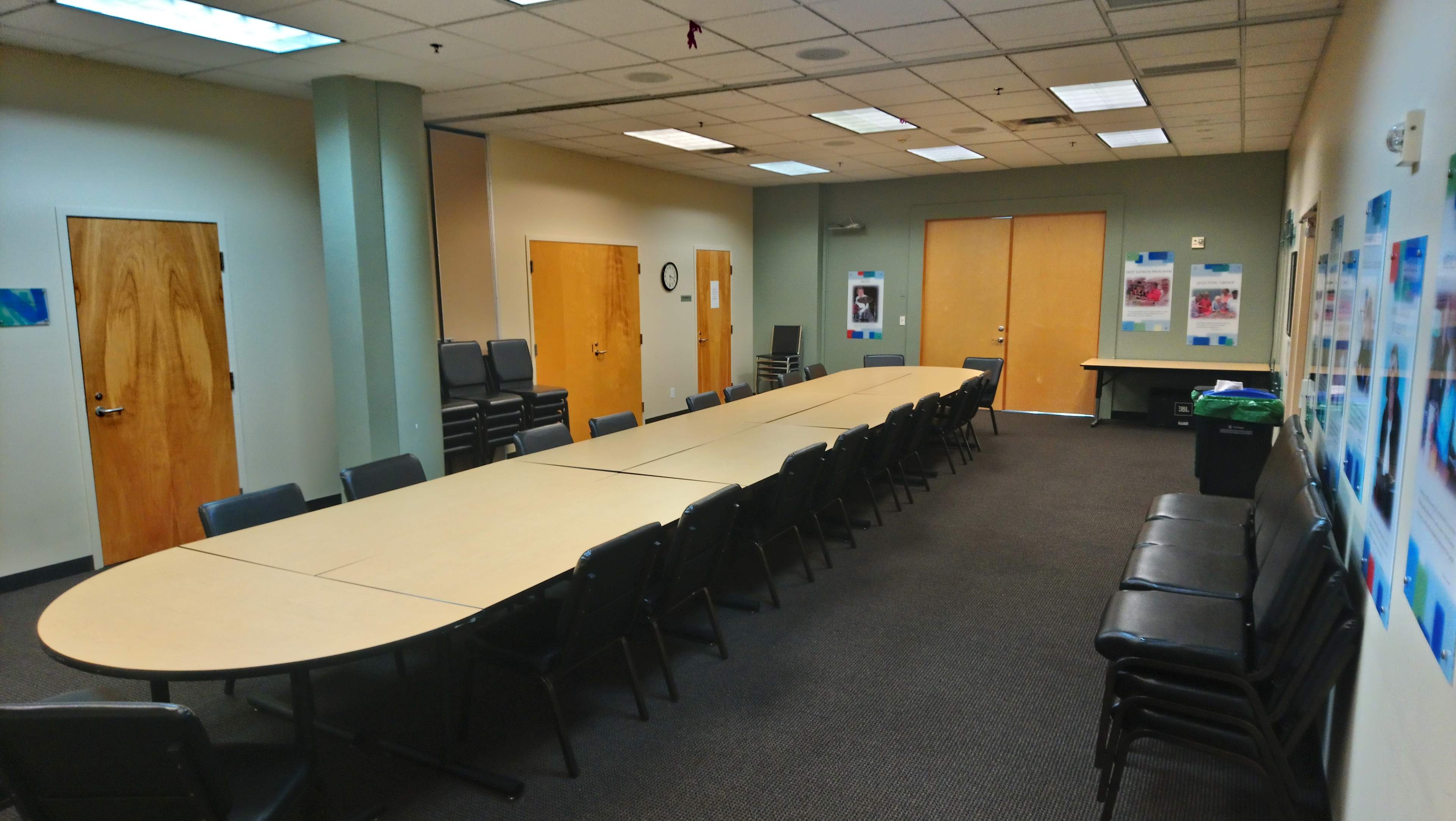 A long, oval conference table is set up in a meeting room with chairs lined along one side and a wall displaying posters.
