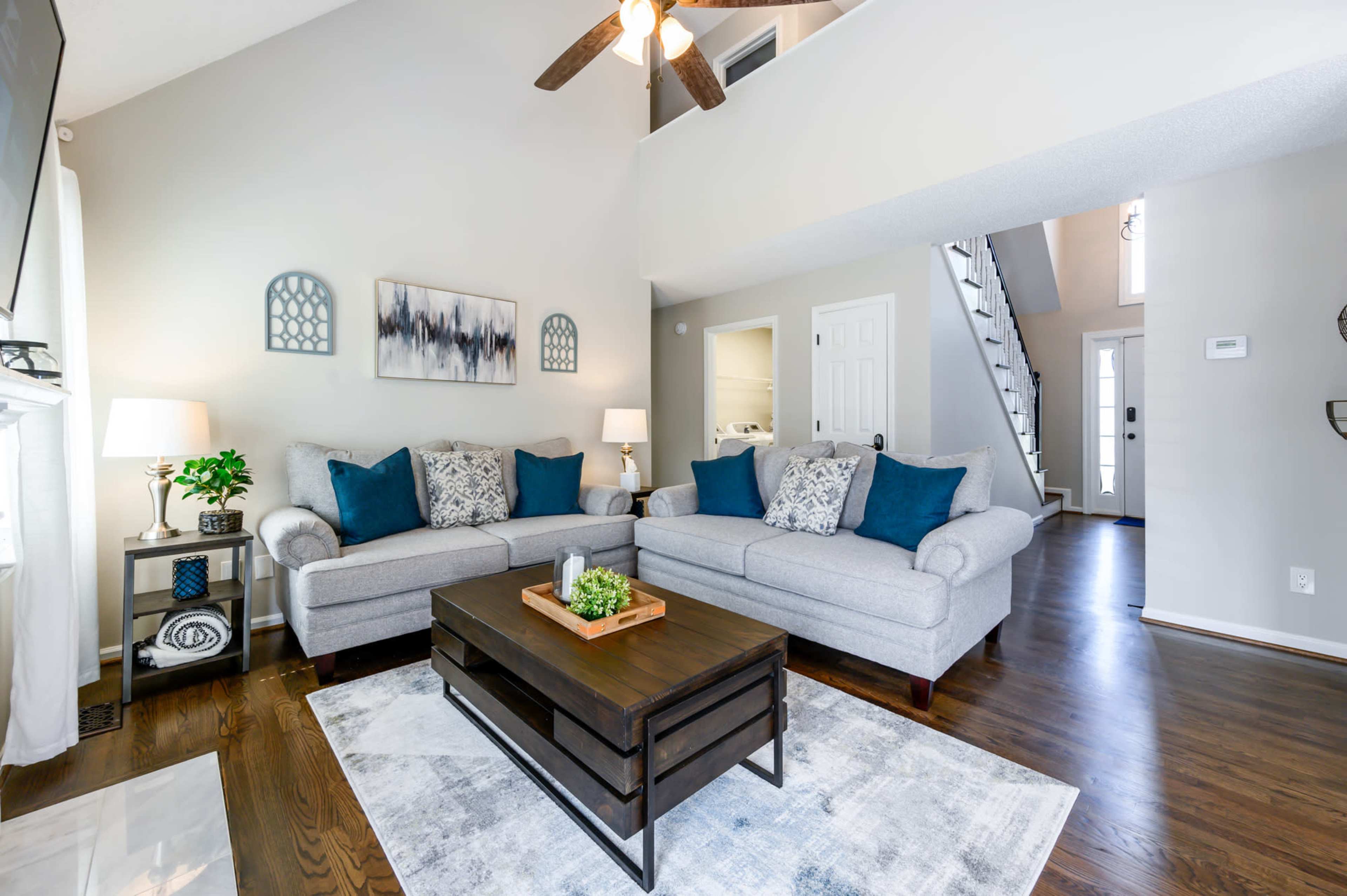 A living room features two gray sofas with blue accent pillows facing a dark wood coffee table on a light area rug, with a staircase and entrance visible in the background.