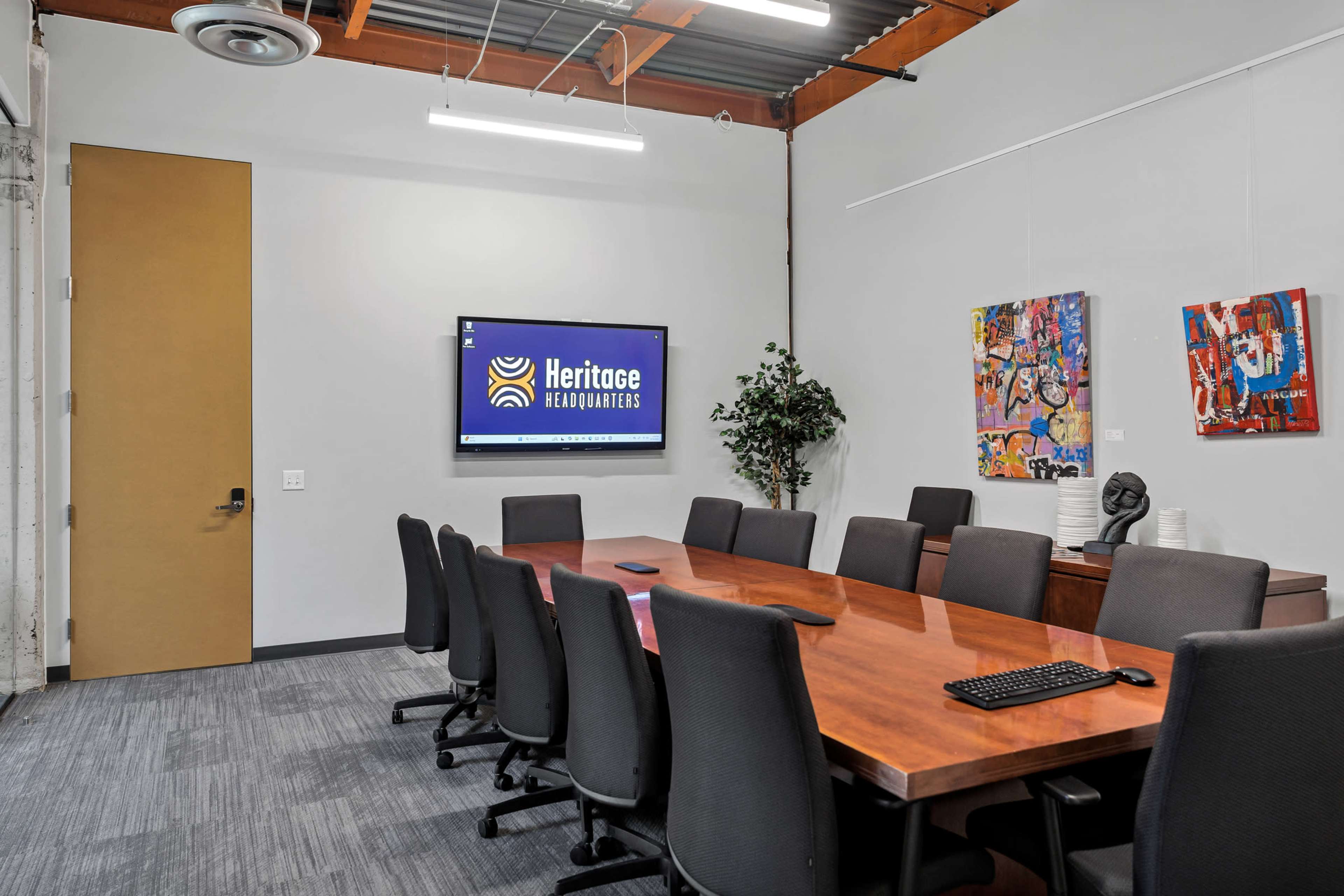 The image shows a modern conference room with a large wooden table, black ergonomic chairs, a wall-mounted screen displaying "Heritage Headquarters," and colorful artwork on the walls.