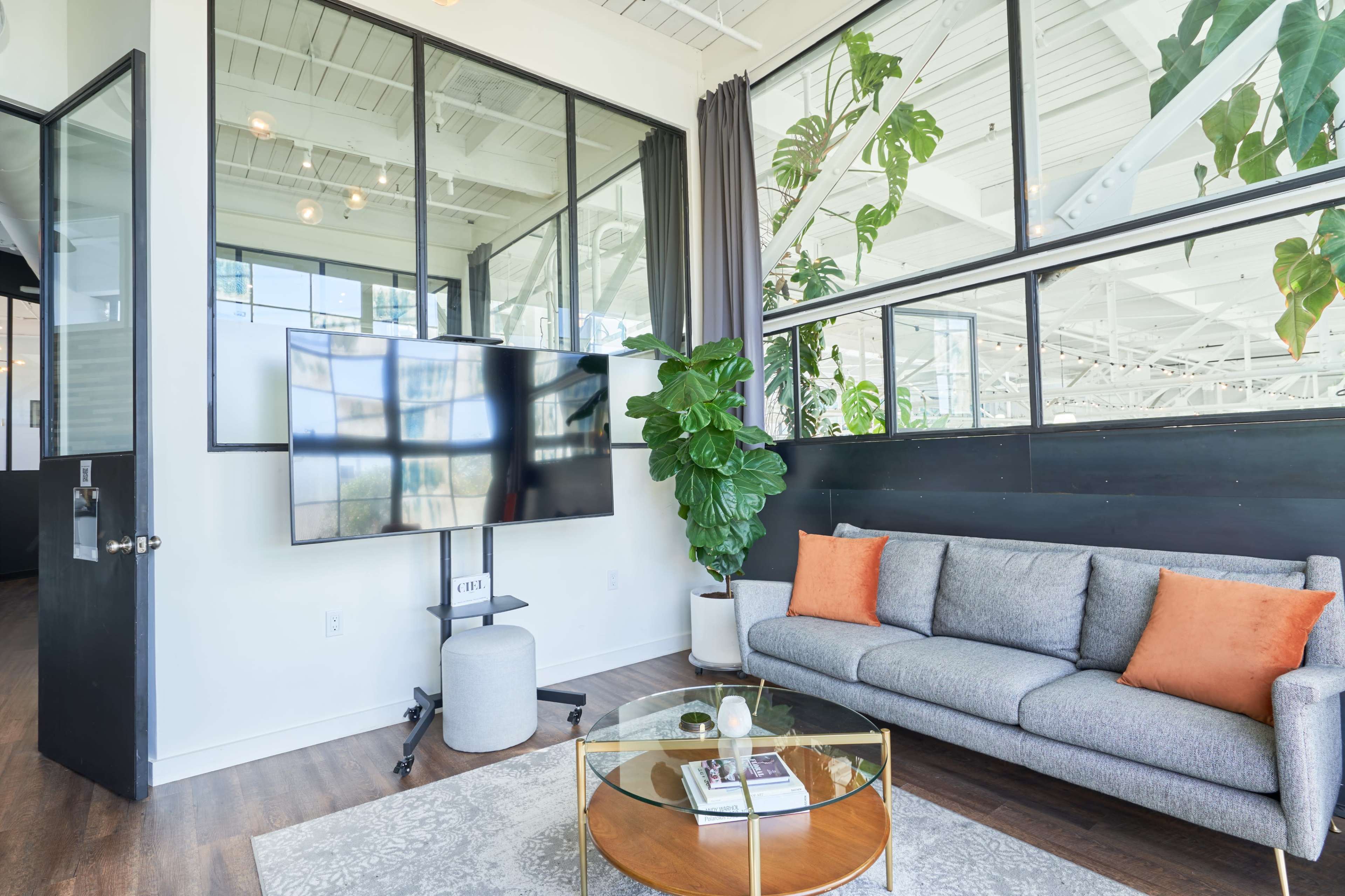 A modern living room with a gray sofa, a round coffee table, a large television mounted on the wall, and large windows that allow natural light to fill the space.