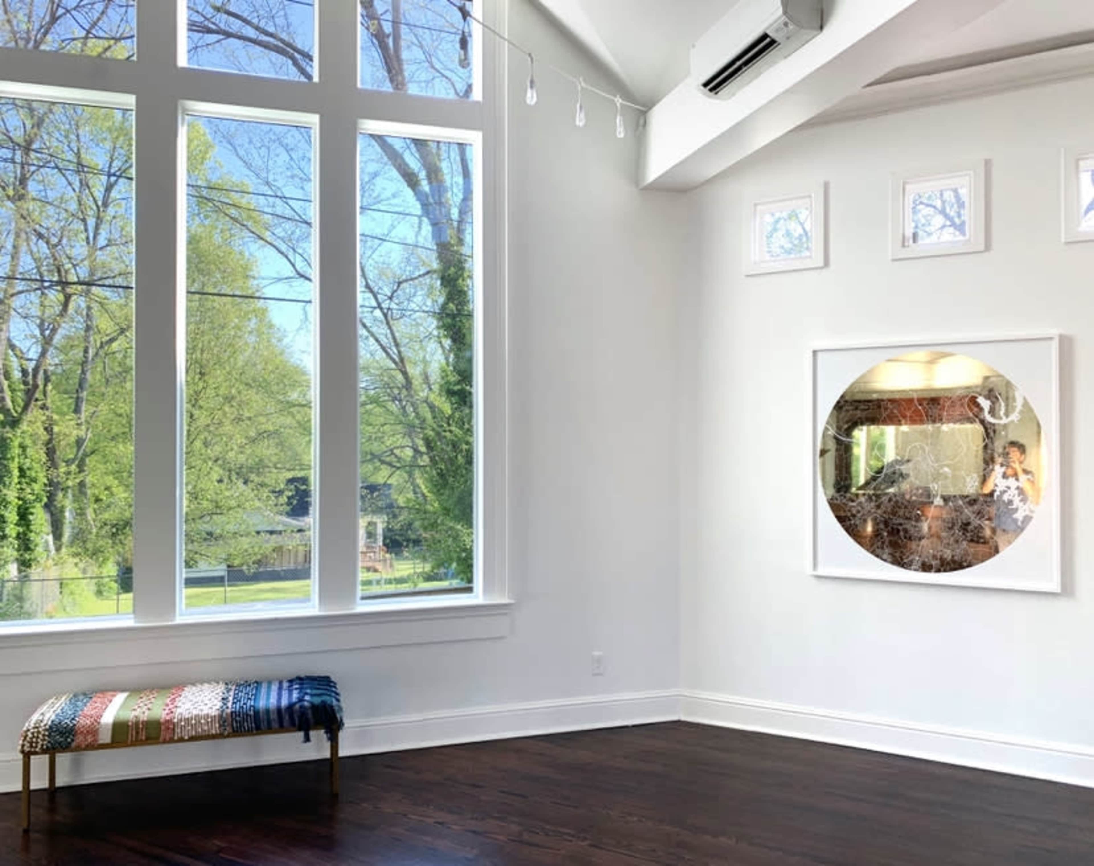 A bright room features large windows overlooking trees, with a bench and a round mirror on the wall.