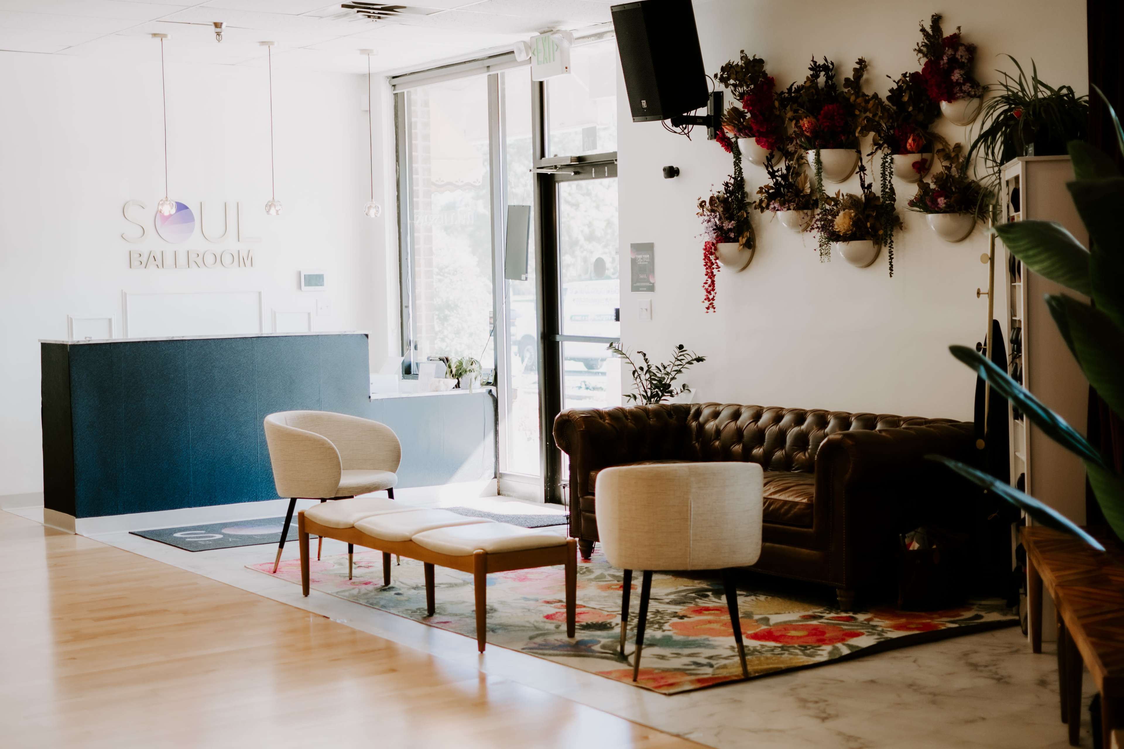 The interior of a dance studio features a reception area with a dark blue front desk and a cozy seating arrangement, including a leather couch and chairs on a decorative rug.