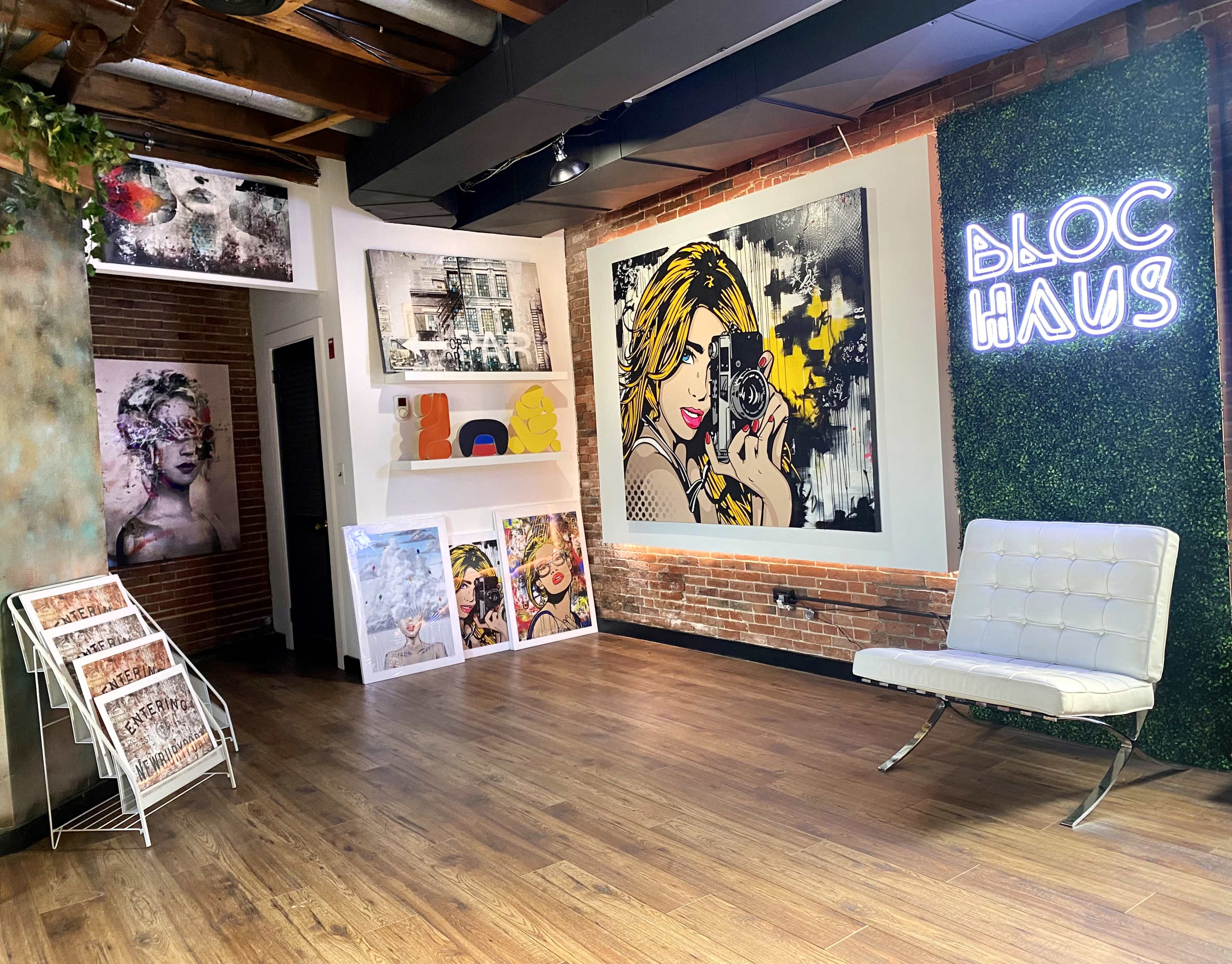 An art gallery space featuring various colorful artworks, including a large portrait of a woman with a camera, and a neon sign that reads "BLOC HAUS" along with a stylish white seat.