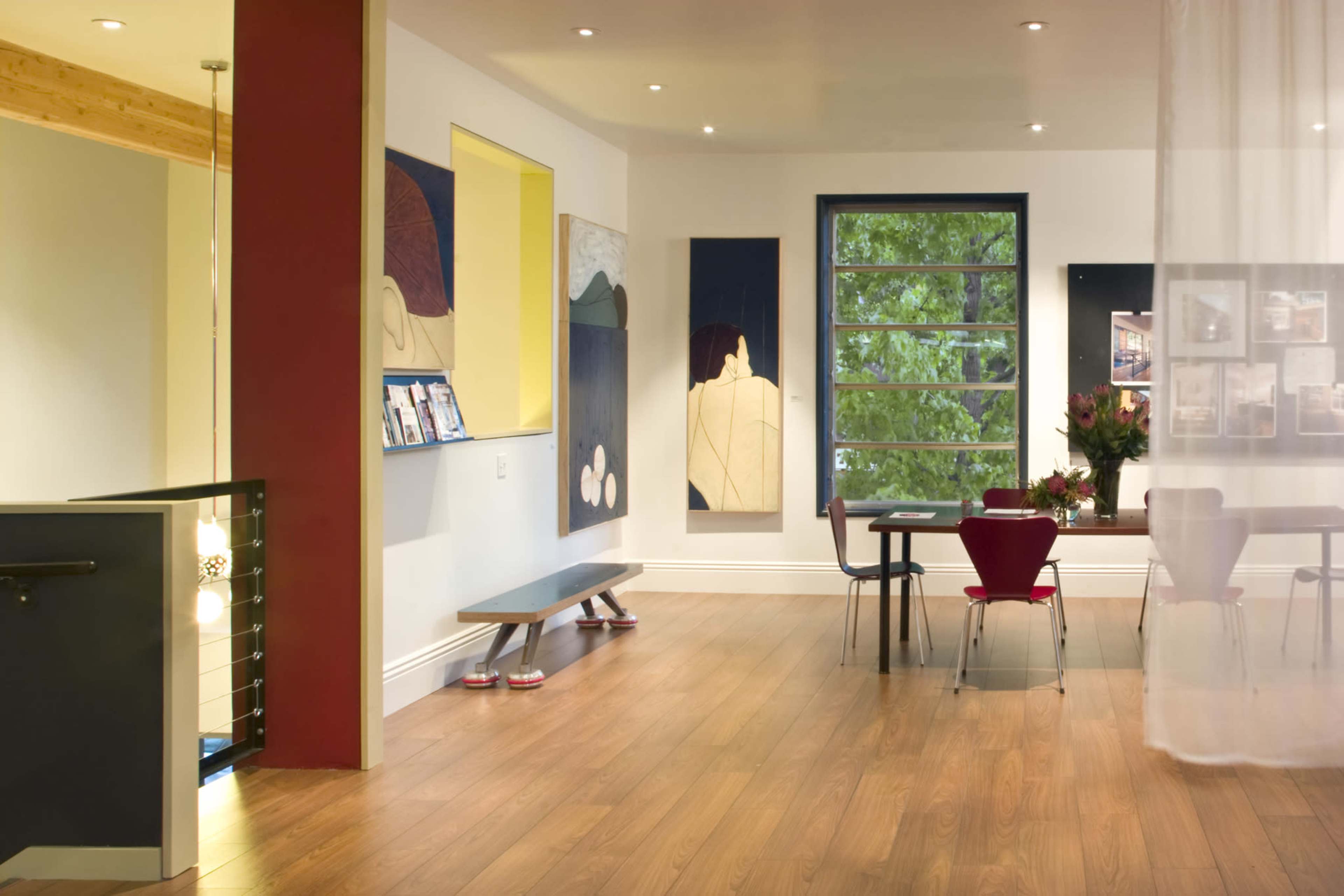 A modern interior space featuring artwork on the walls, wooden flooring, and a dining area with a table and red chairs near a large window.