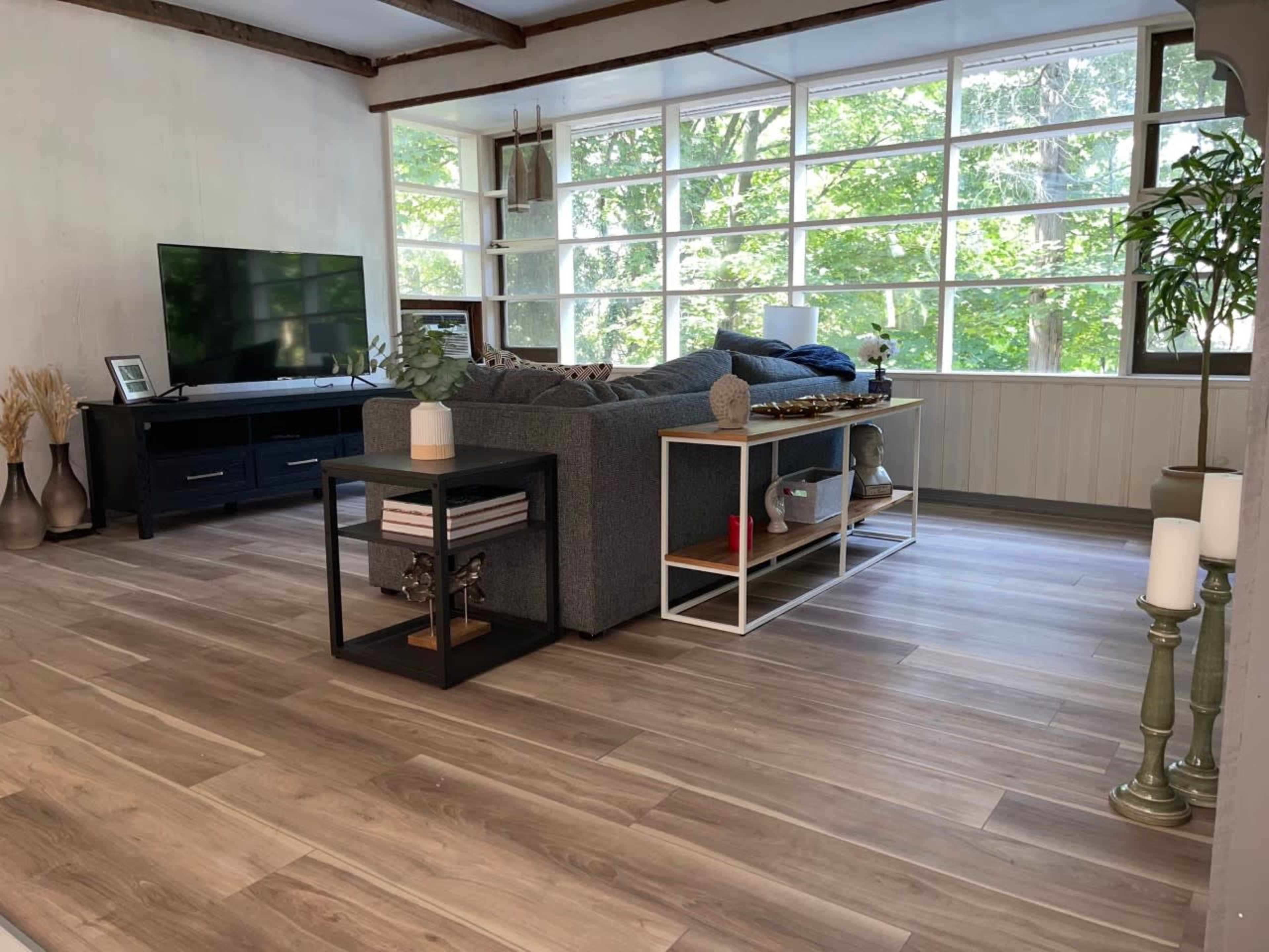 A modern living room with a gray sofa, a television on a dark console, and large windows letting in natural light, surrounded by green trees outside.