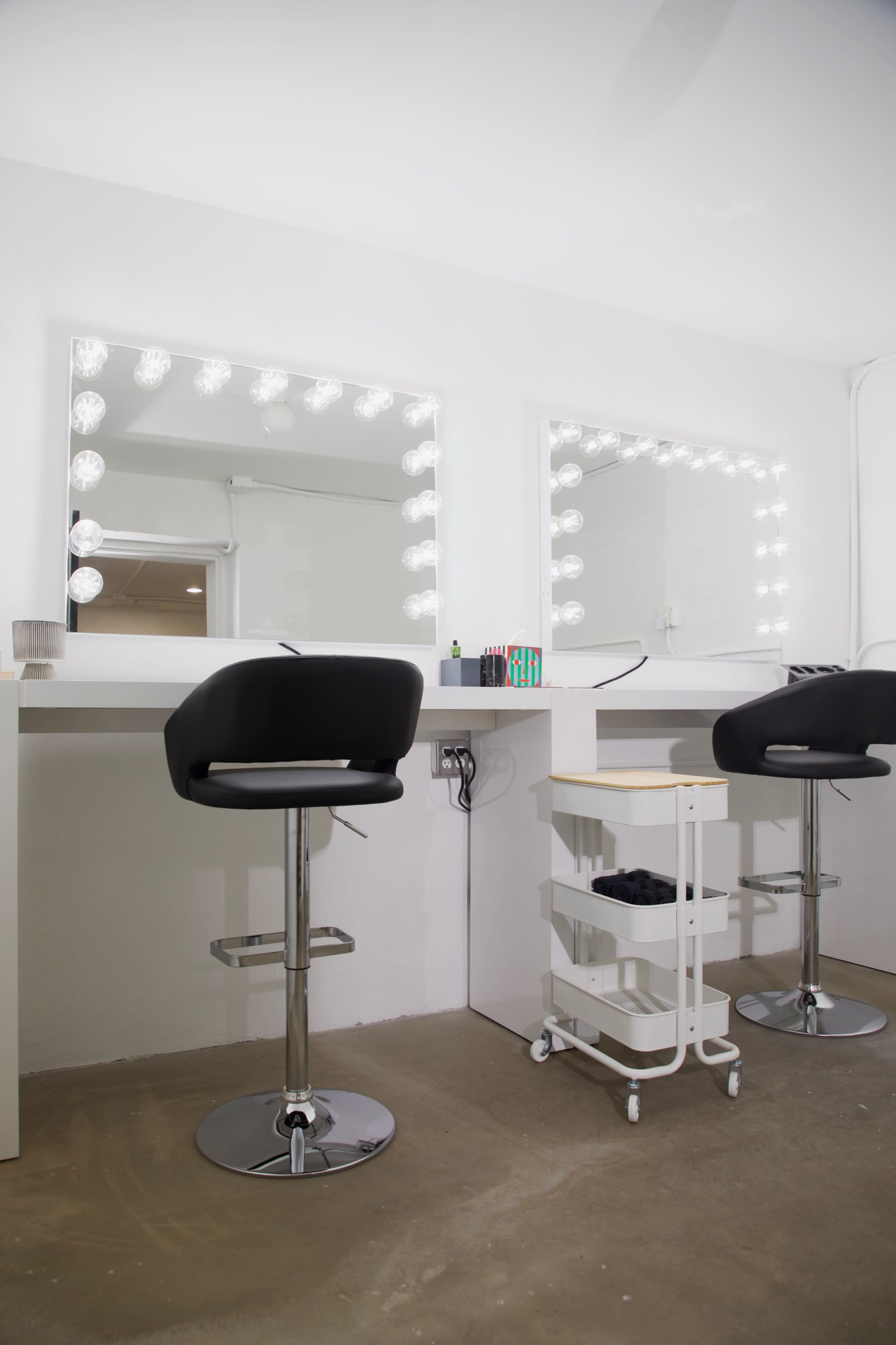 The image shows a minimalist makeup station with two high stools, illuminated mirrors, and a small cart for storage.