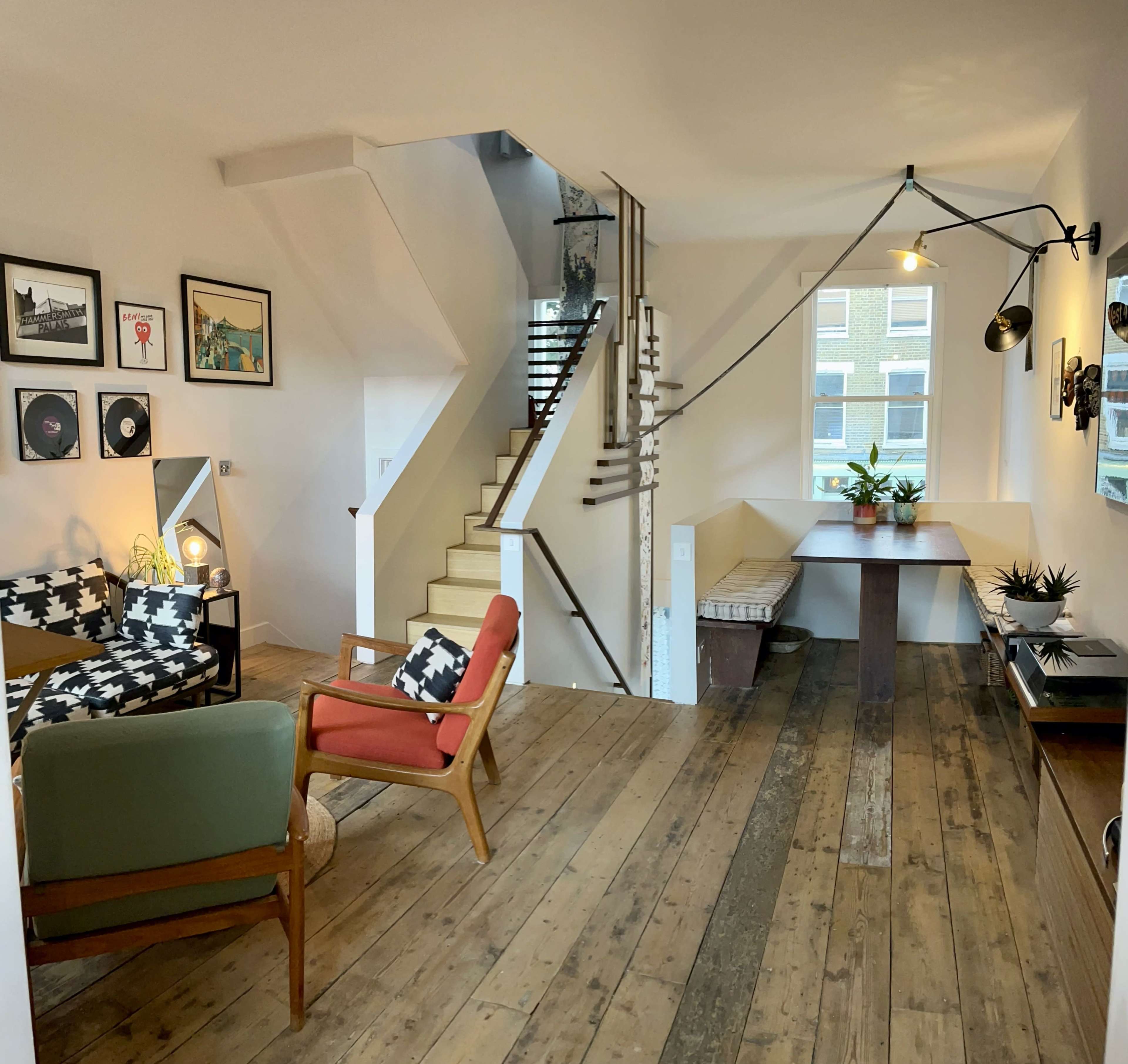 The image shows a cozy living space with a dining area, a staircase, and artwork on the walls, featuring wooden floors and various seating options.