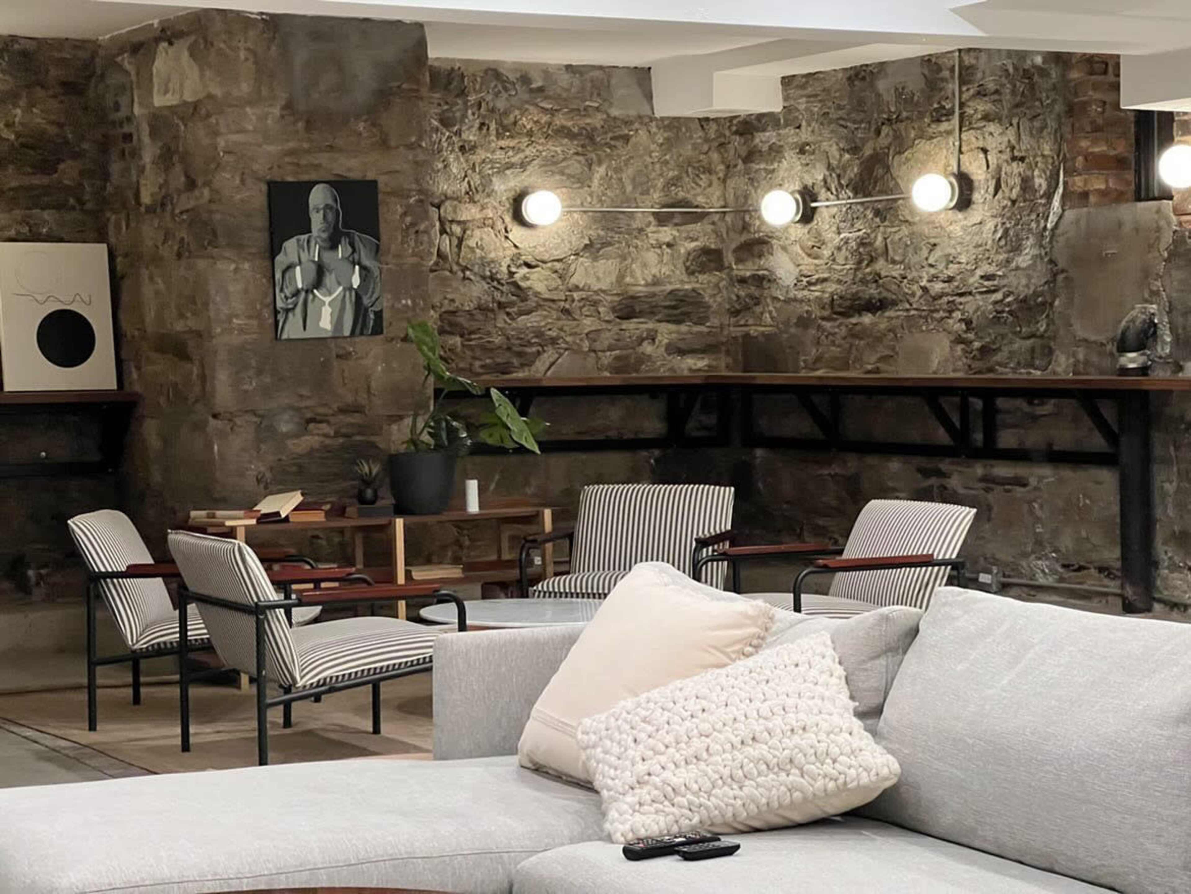 A stylish lounge area with a sectional sofa in the foreground, accompanied by a few striped armchairs and wooden tables against a wall of exposed stone.
