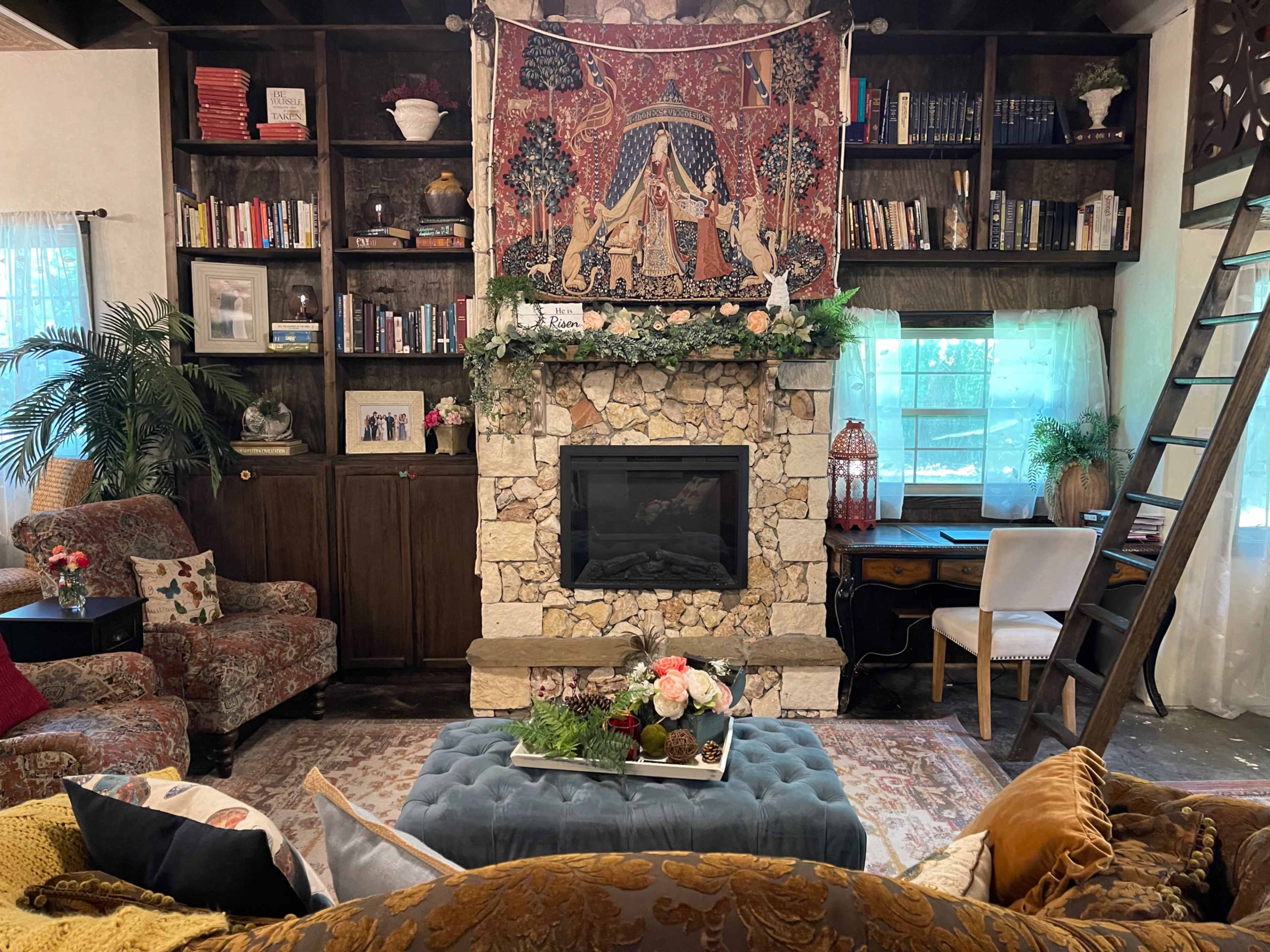 The image shows a cozy living room with a stone fireplace, bookshelves, and a decorative tapestry above the mantel.