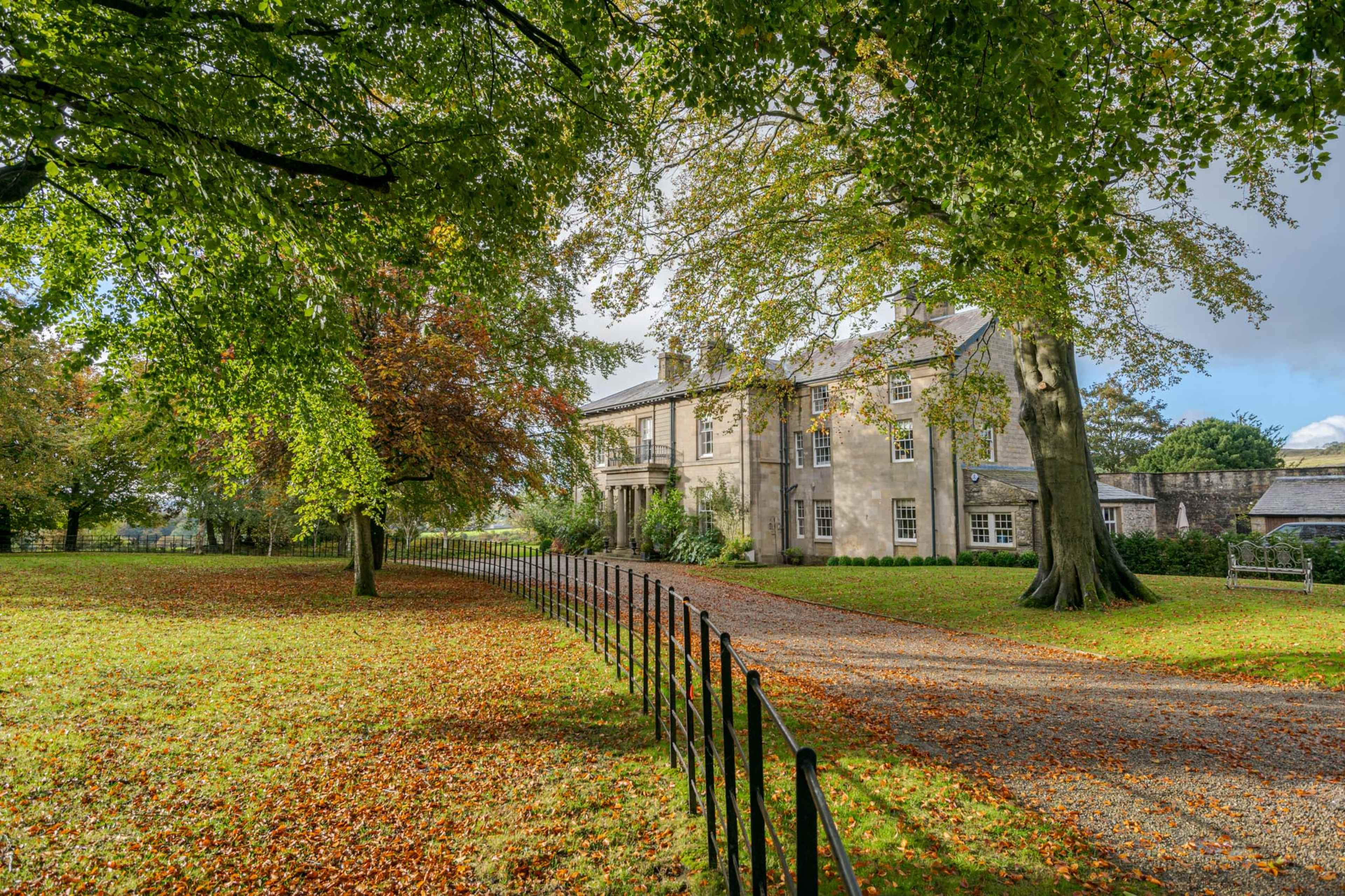 A large stone house with a gravel driveway is surrounded by trees and autumn leaves.
