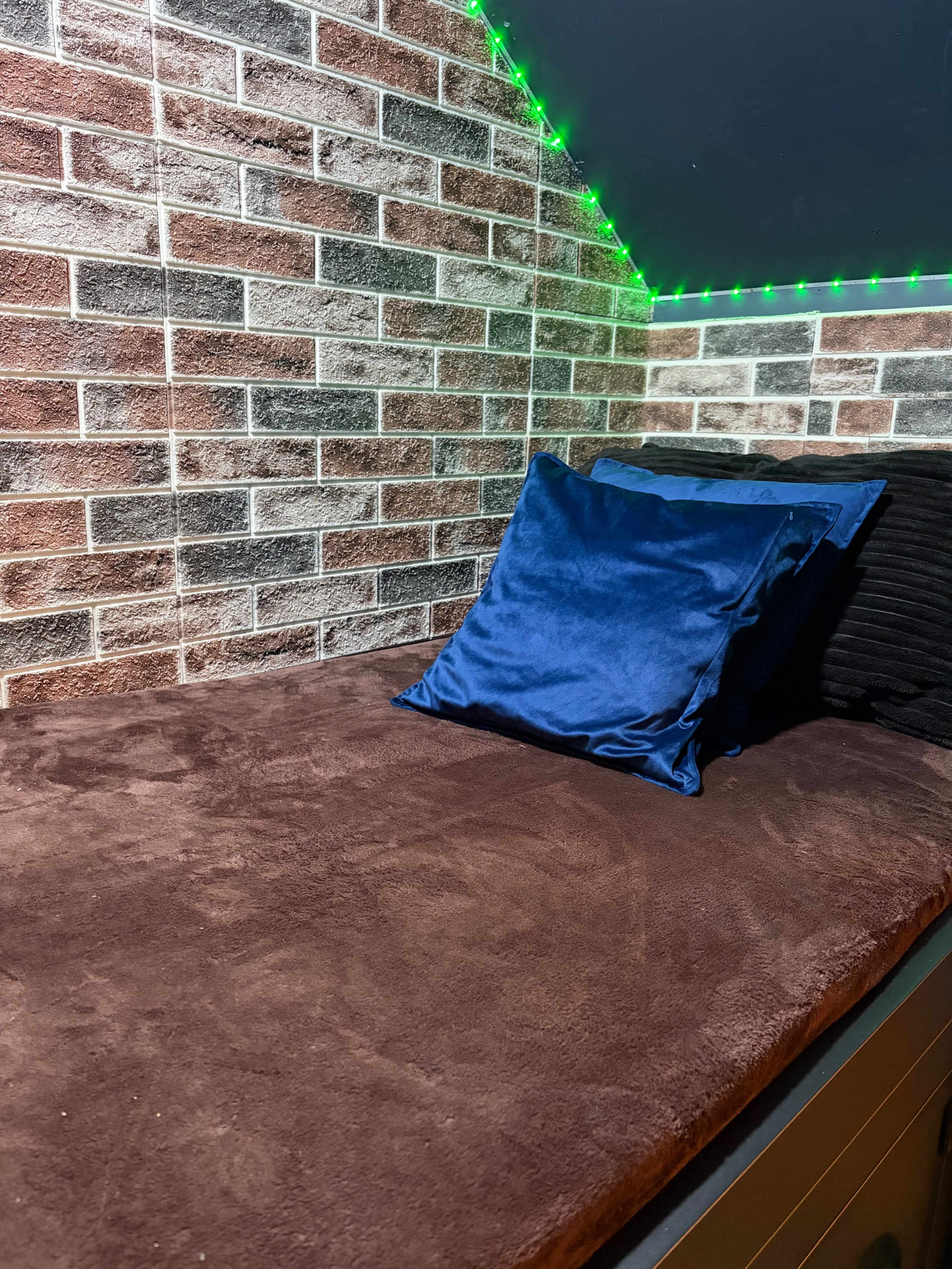 The image shows a cozy nook with a brown plush surface, a blue pillow, and a brick-patterned wall, illuminated by green LED lights.