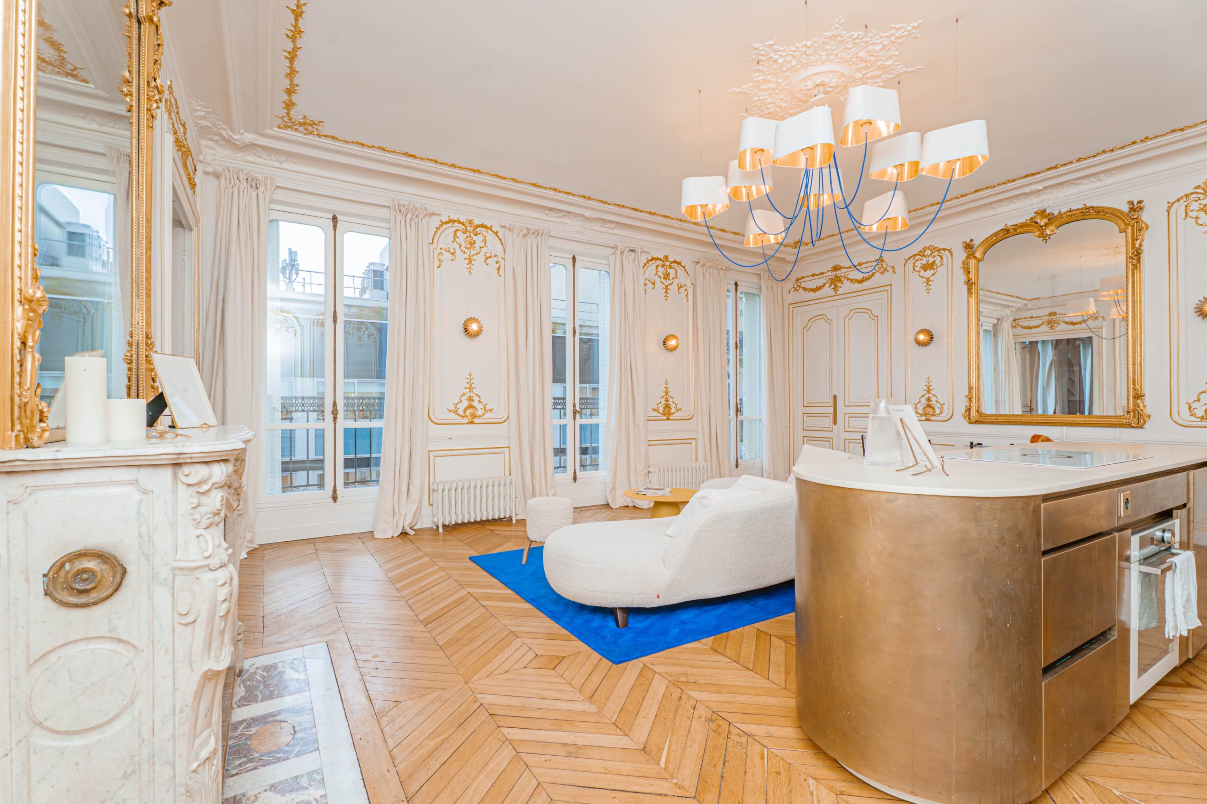 The room features elegant gold accents, a large circular chandelier, and a cozy white sofa on a blue rug, with large windows allowing natural light to fill the space.