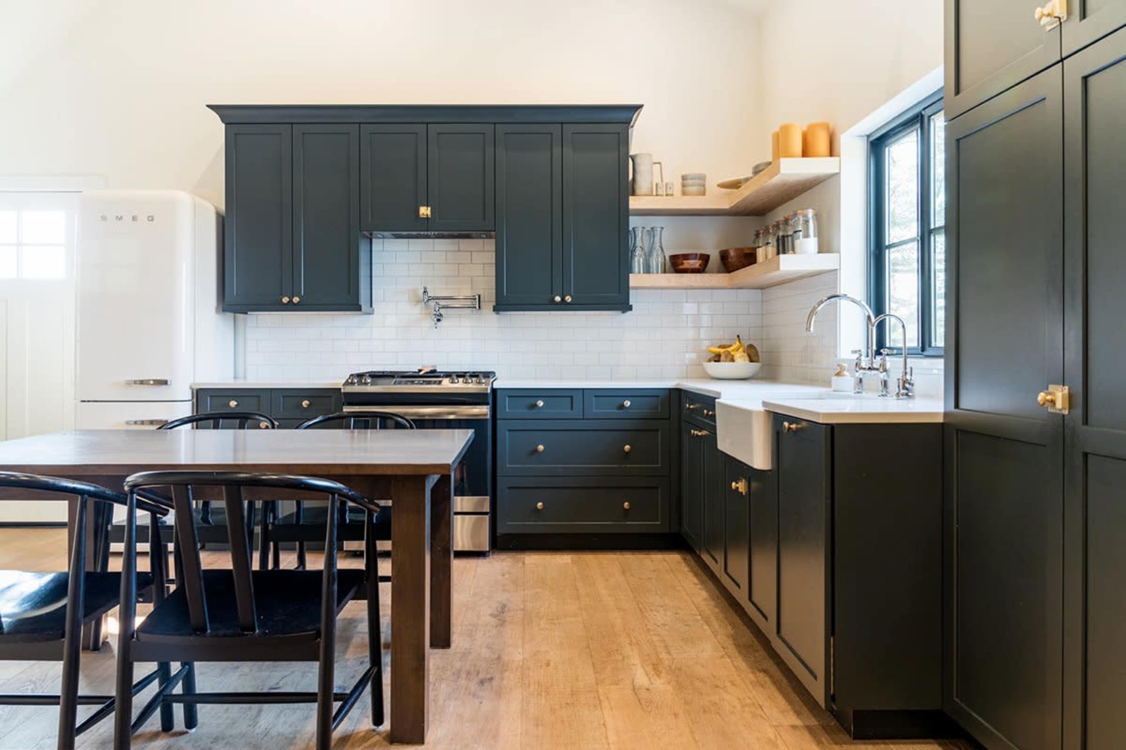 The image shows a modern kitchen featuring dark green cabinets, white countertops, a stainless steel stove, and a wooden dining table surrounded by black chairs.