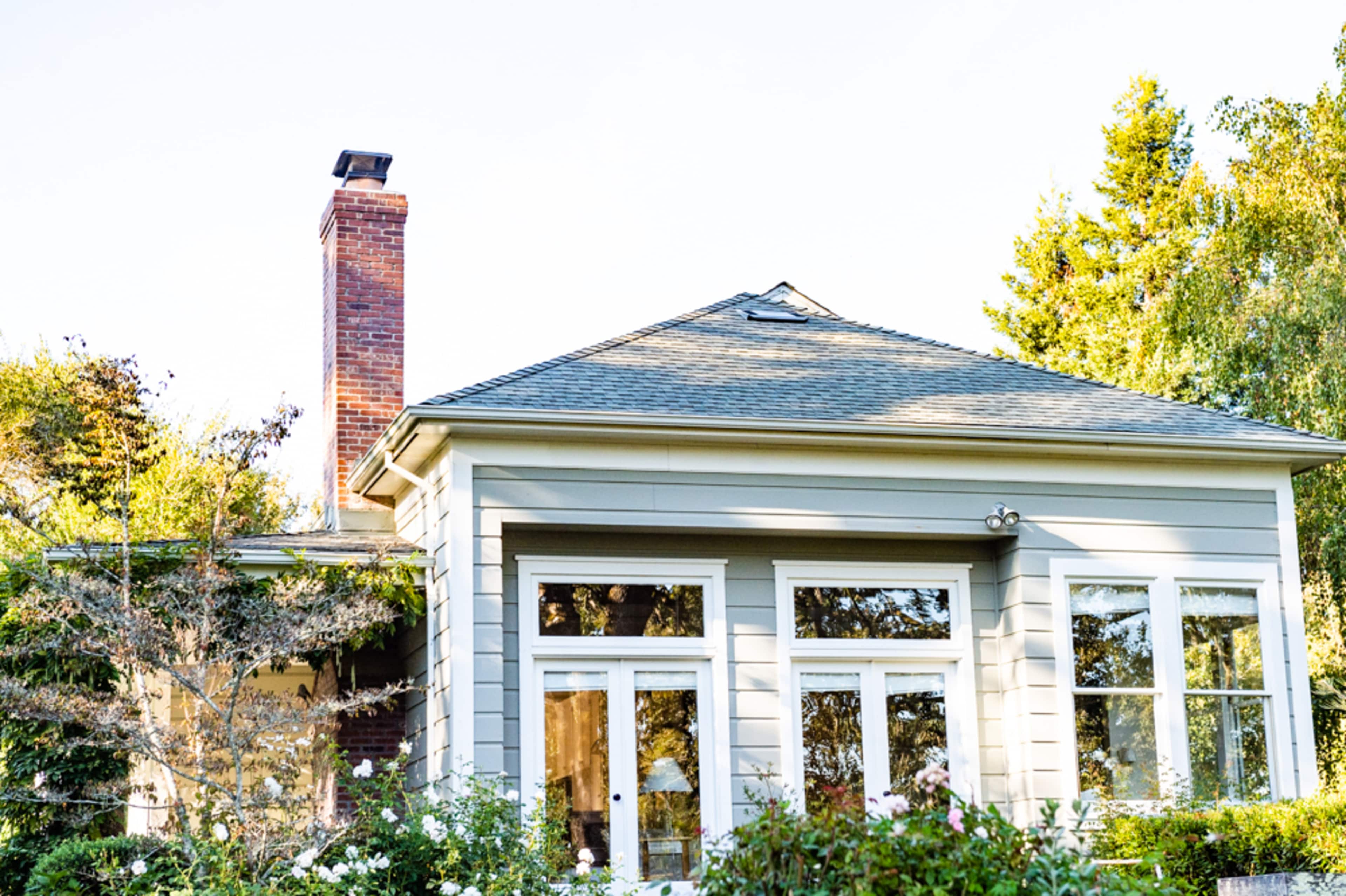 A single-story house with a chimney and large windows is surrounded by trees and a garden.
